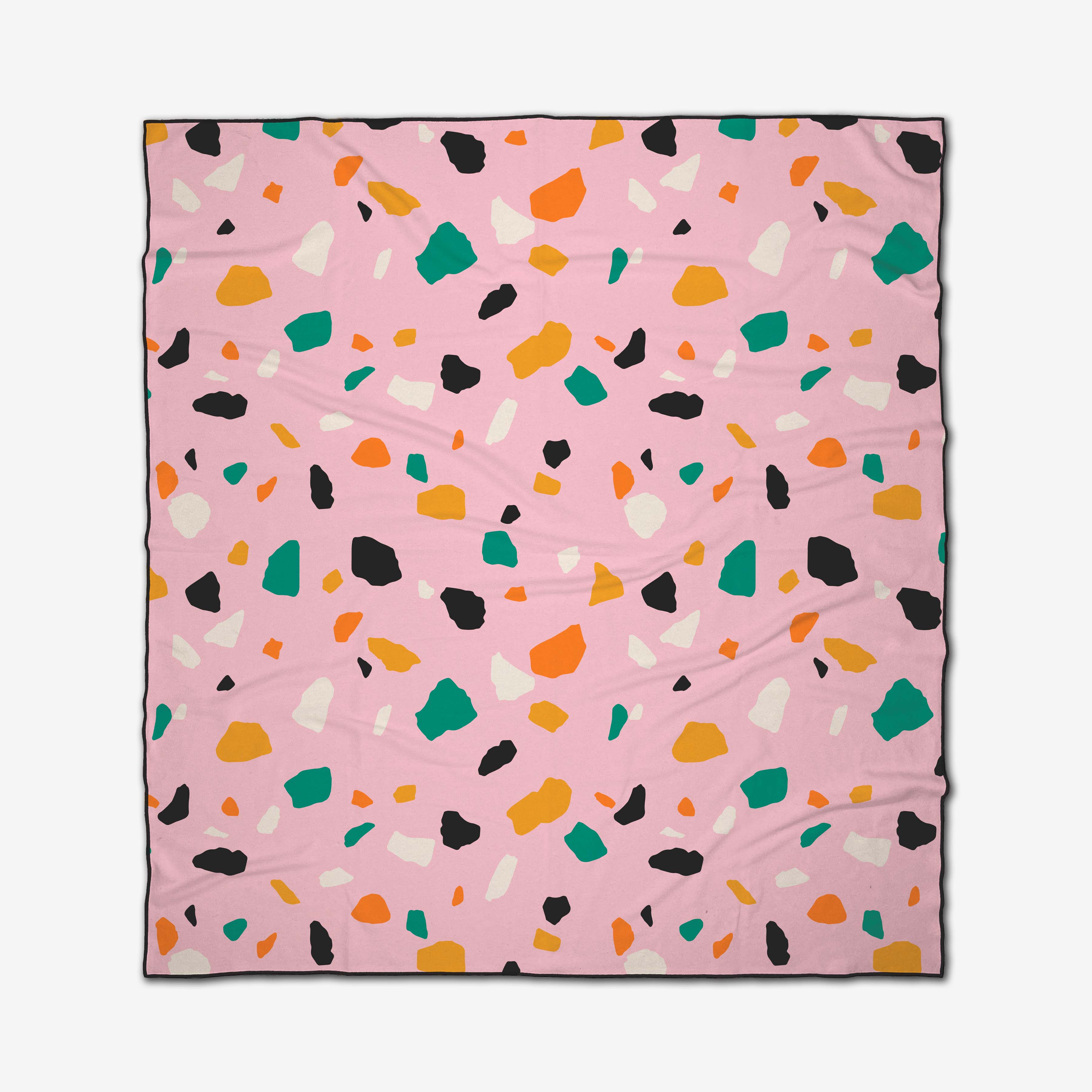 The Megan Roy Pink Terrazzo beach blanket features a pink background with abstract black, white, orange, yellow, and green shapes. Square-shaped and made from quick-drying recycled materials for stylish sustainability.