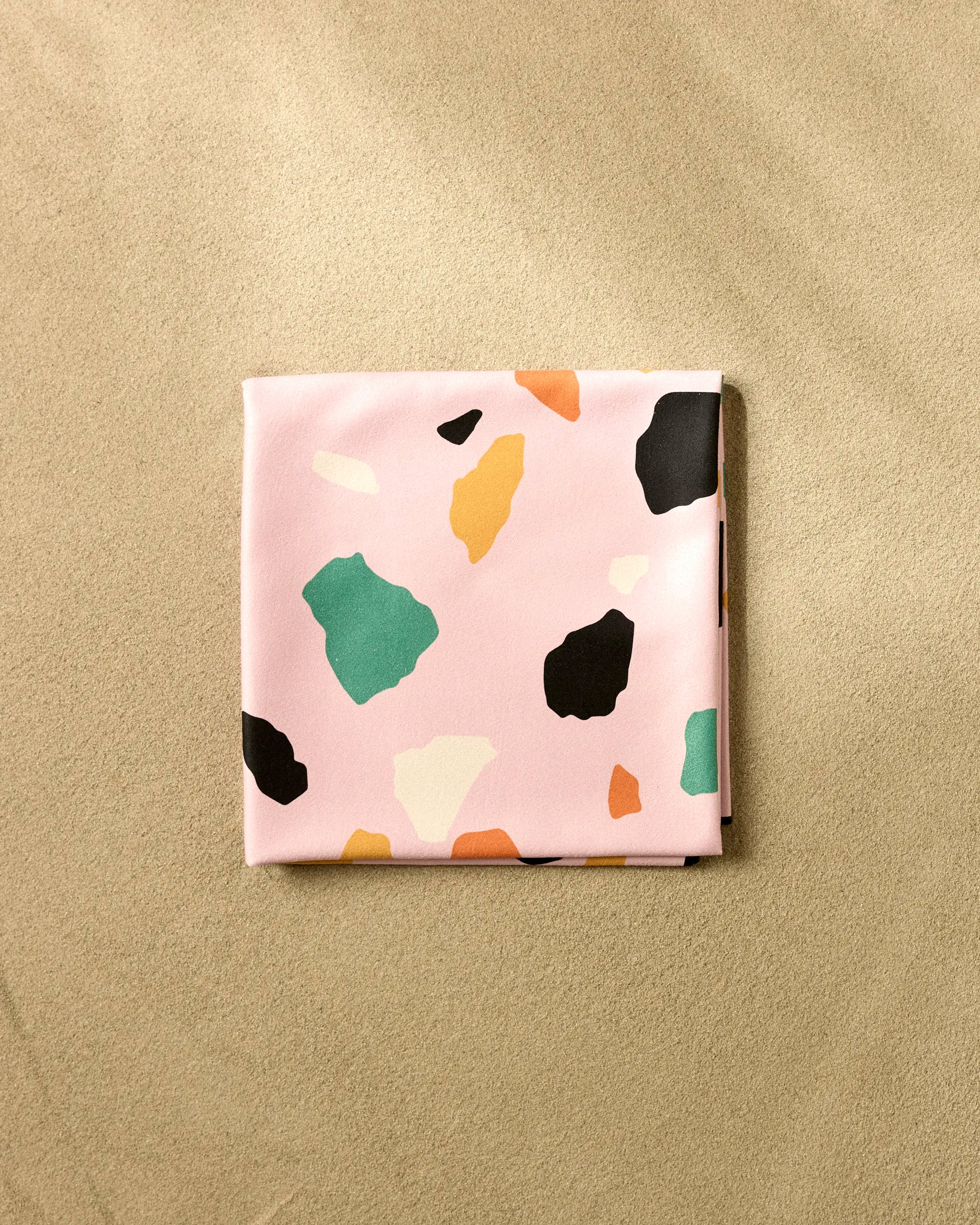 The Megan Roy Pink Terrazzo beach blanket, made from quick-drying recycled materials, features a pink background with colorful abstract shapes and is shown folded on a beige textured surface.