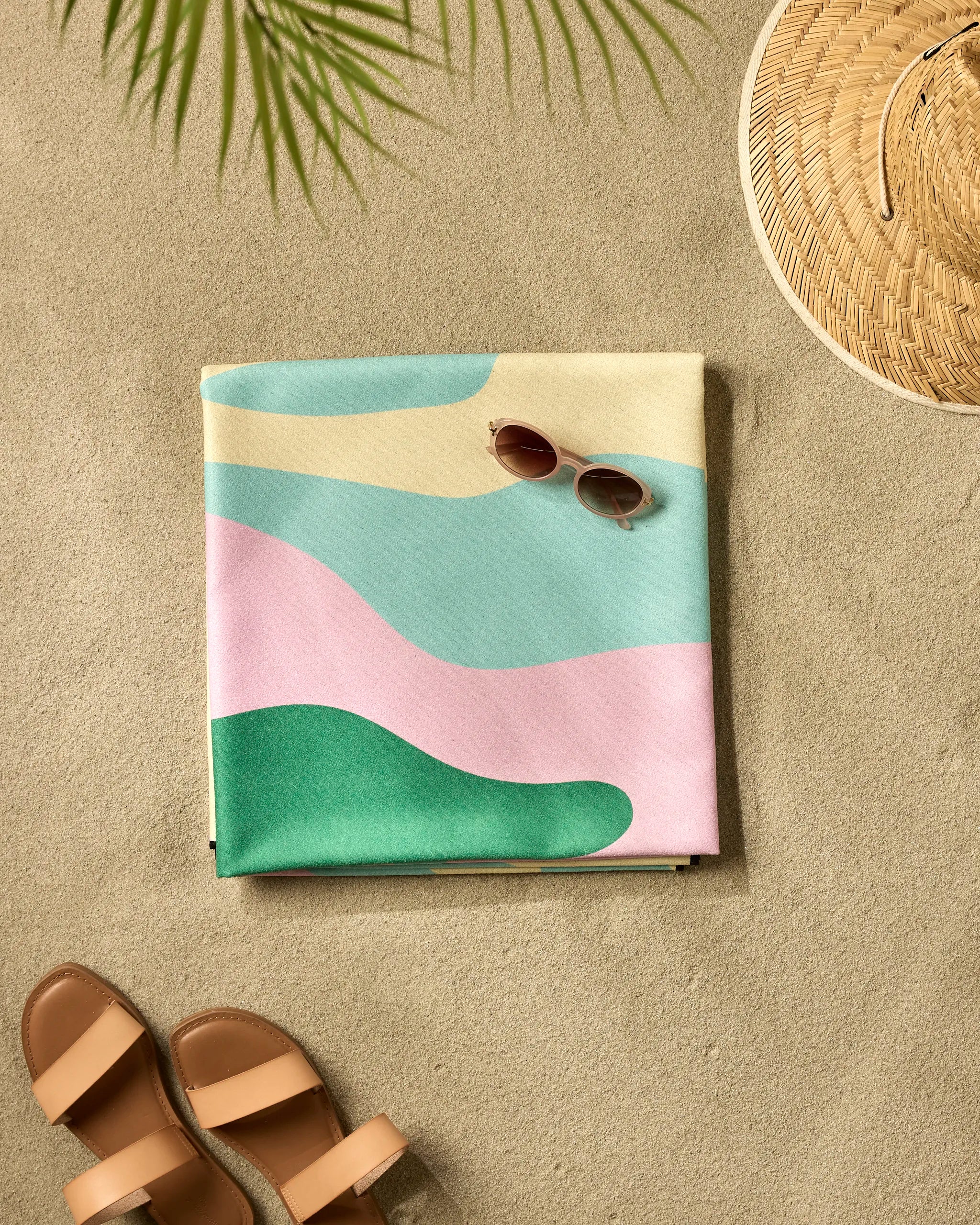 The GEOMETRY "Pinks And Greens" recycled beach blanket, featuring abstract pastel patterns, is folded on sandy ground beside sunglasses, sandals, and a straw hat under a palm leaf’s shadow.