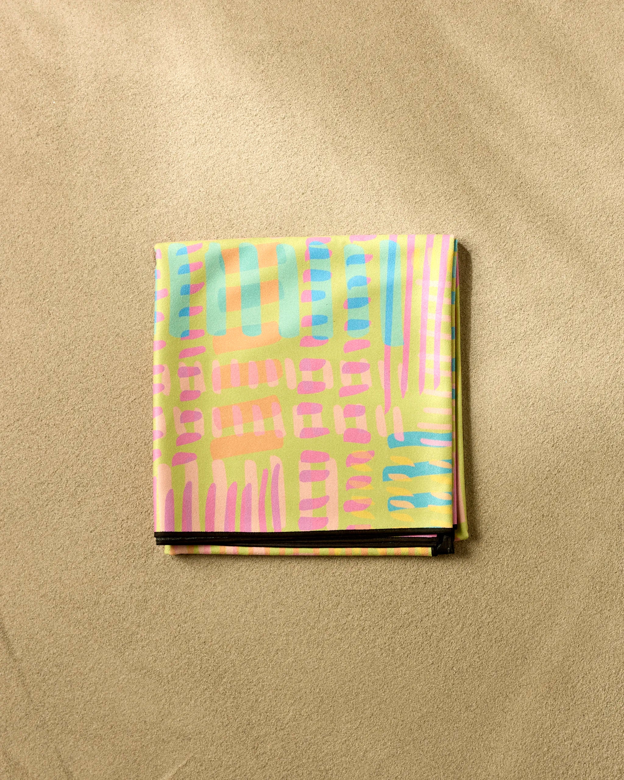The "Positive Vibes" beach blanket by Sareka Unique, featuring a pastel geometric pattern in pink, blue, green, and yellow, is folded neatly on a beige textured surface.