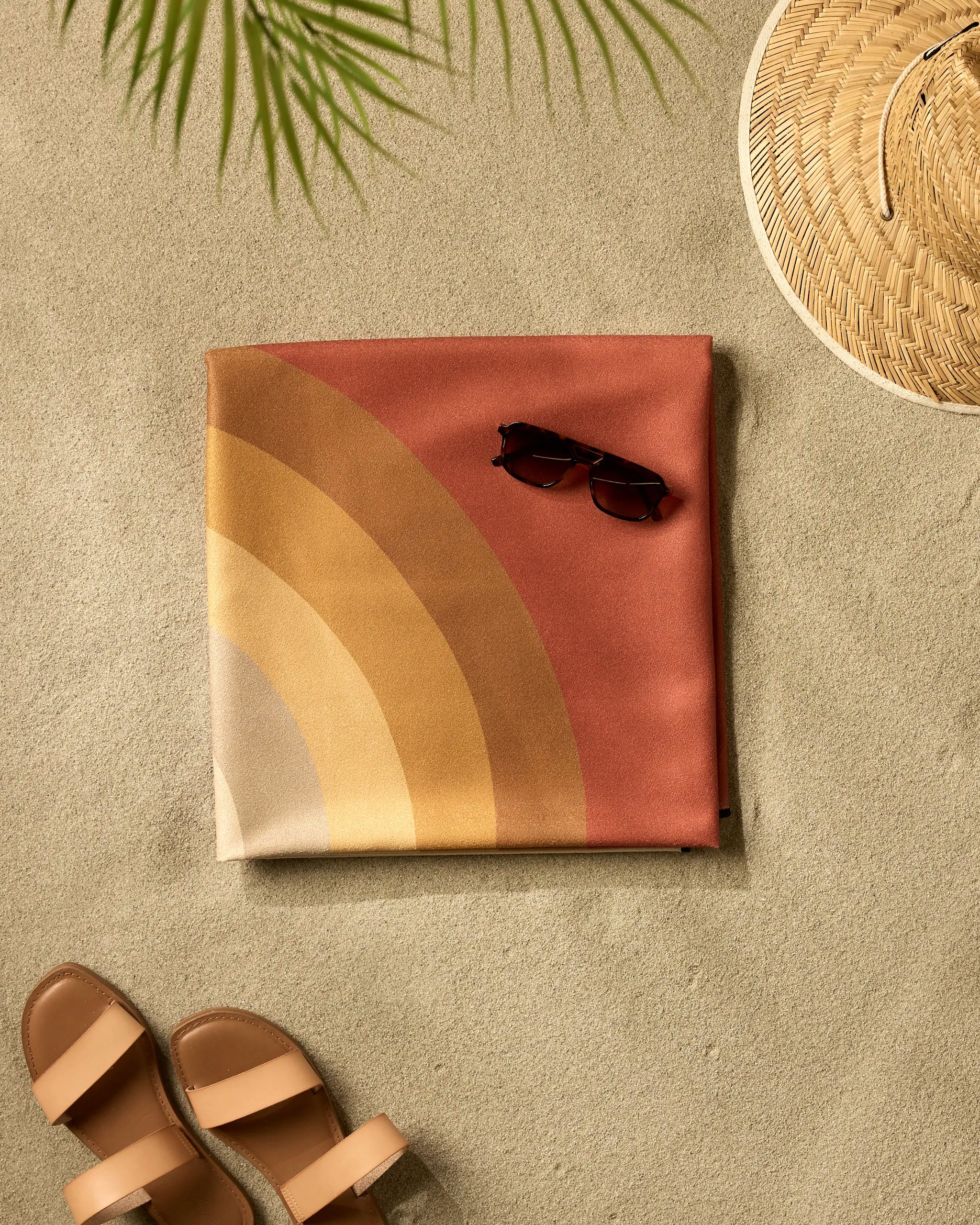 The GEOMETRY "Pumpkin Spice" quick-drying towel, with warm rainbow stripes, is arranged on the sand beside brown sunglasses, a woven straw hat, tan sandals, and topped with a green palm frond.