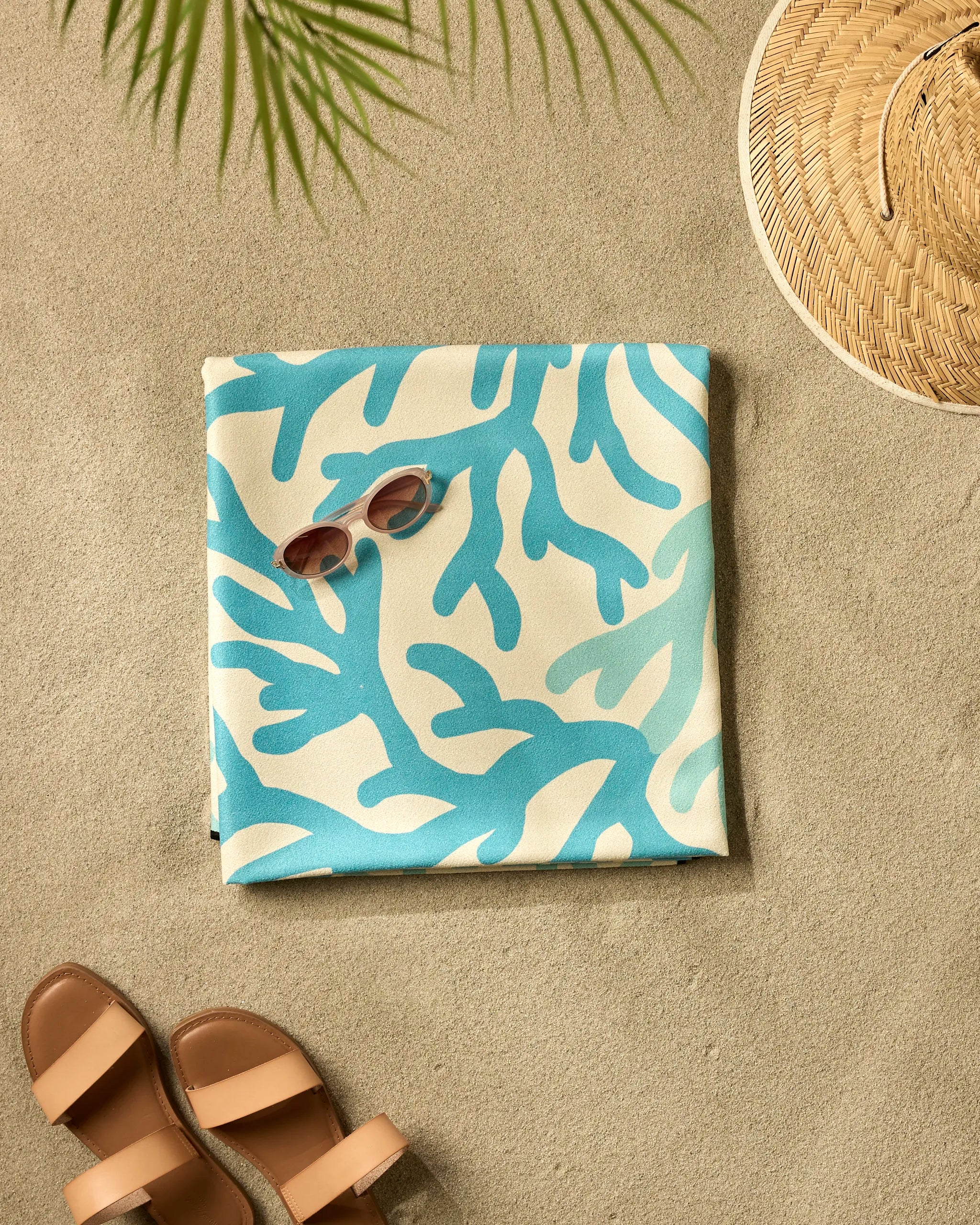 A pair of sunglasses rests on a folded GEOMETRY Sea Corals Beach Blanket with a blue-and-white coral pattern on the sand, beside brown sandals, a straw hat, and a palm leaf casting its shadow.