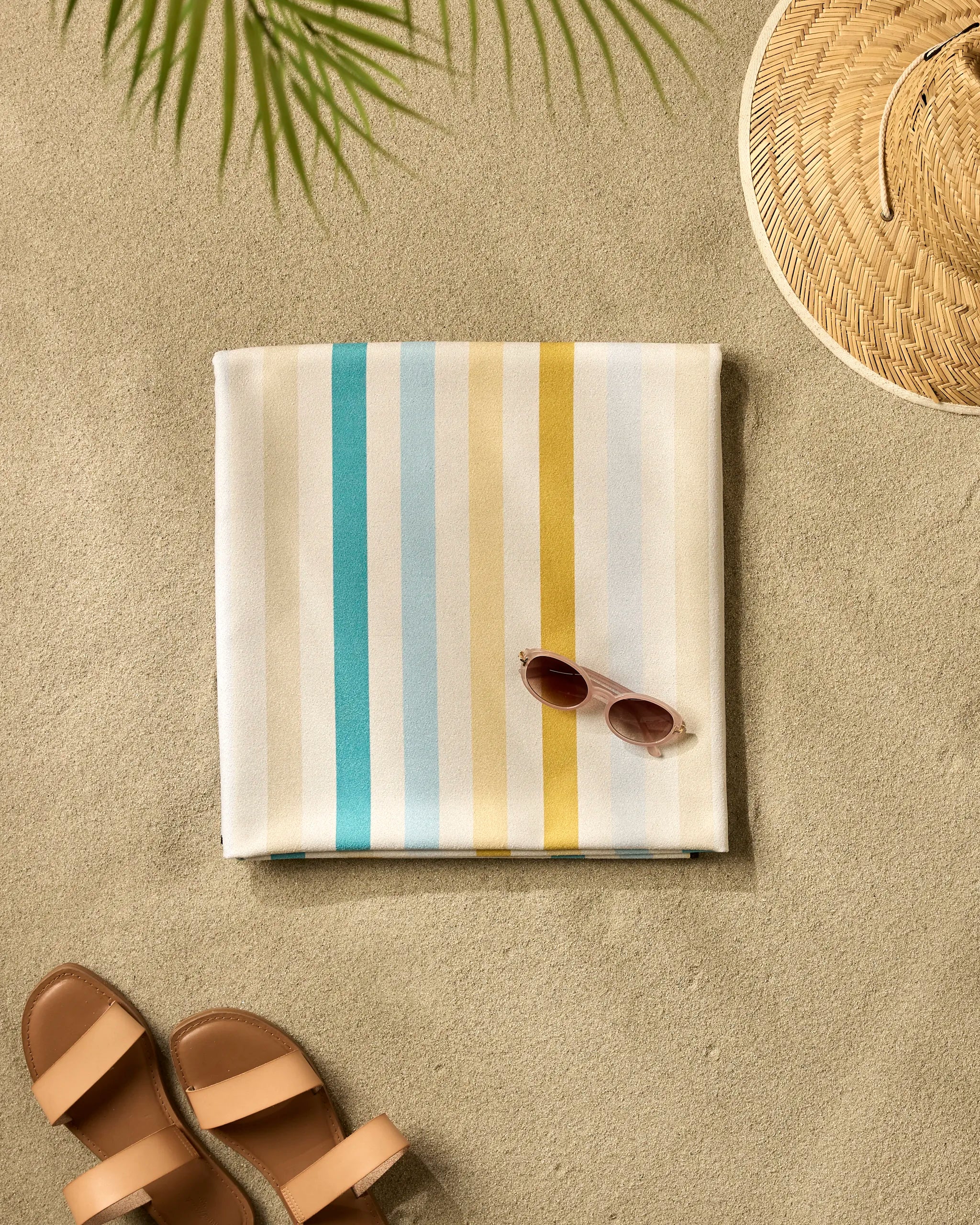 The GEOMETRY Seaside Sunshine quick-dry beach blanket is folded on sand with sunglasses on top, beside a straw hat, brown sandals, and a palm leaf casting a shadow.
