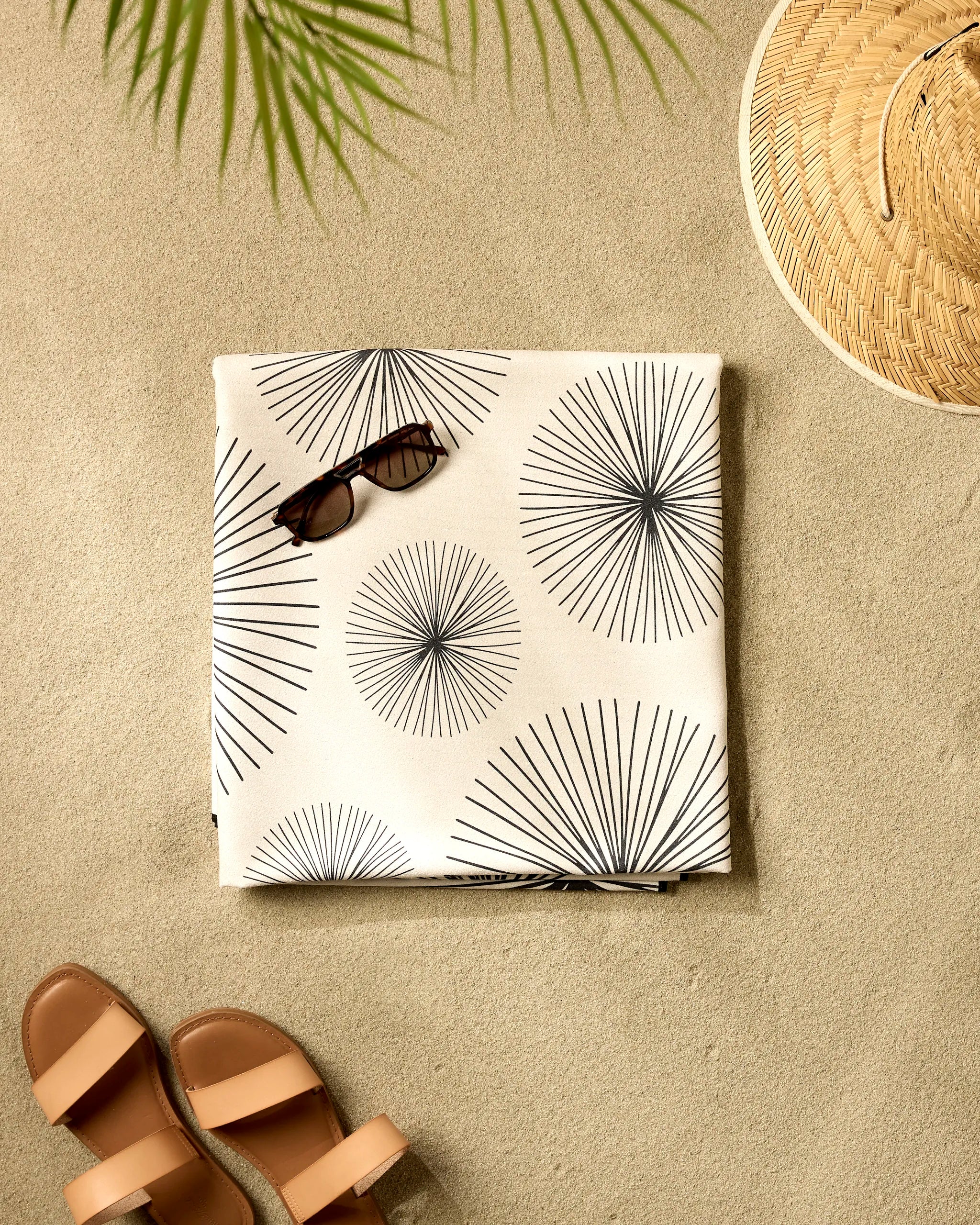 The GEOMETRY Sky Party set features sandals, sunglasses, a straw hat, and a folded quick-dry recycled beach towel with a black sunburst pattern, laid on sand with palm leaf shadows in the corner.