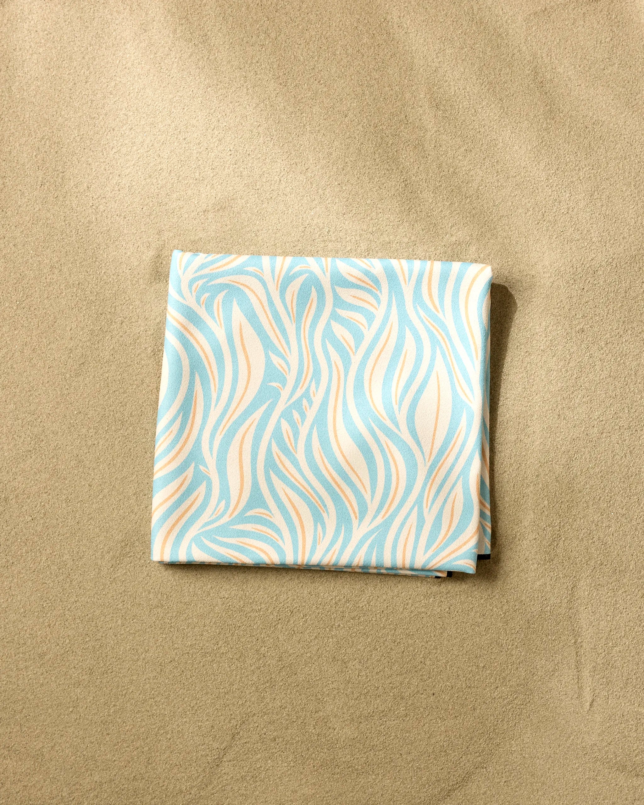 The Rachel Parker Sky Willow Wave, a quick-drying beach blanket made from recycled materials with a light blue and beige wavy pattern, is folded on smooth sand.