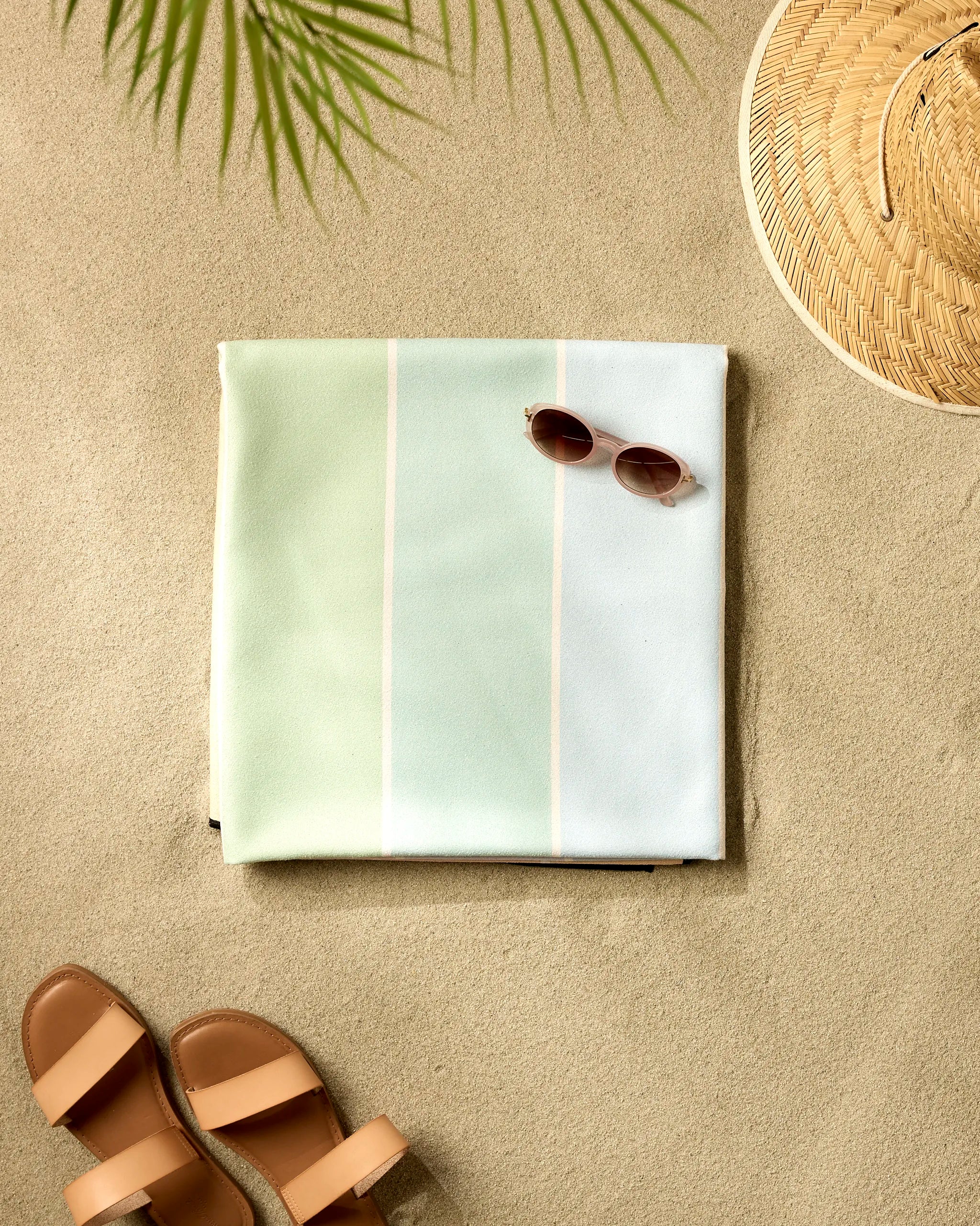 A GEOMETRY Smarties pastel beach blanket is folded on sandy ground, paired with sunglasses, a straw hat, and brown sandals under the shadow of a palm leaf.