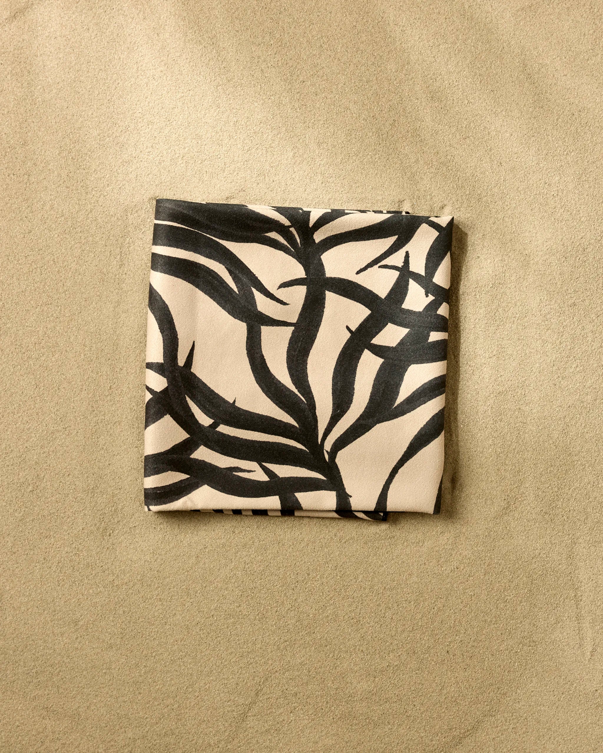 The Geometry Solar Flare beach blanket, folded and beige with a bold black abstract leaf pattern, rests on the sand.
