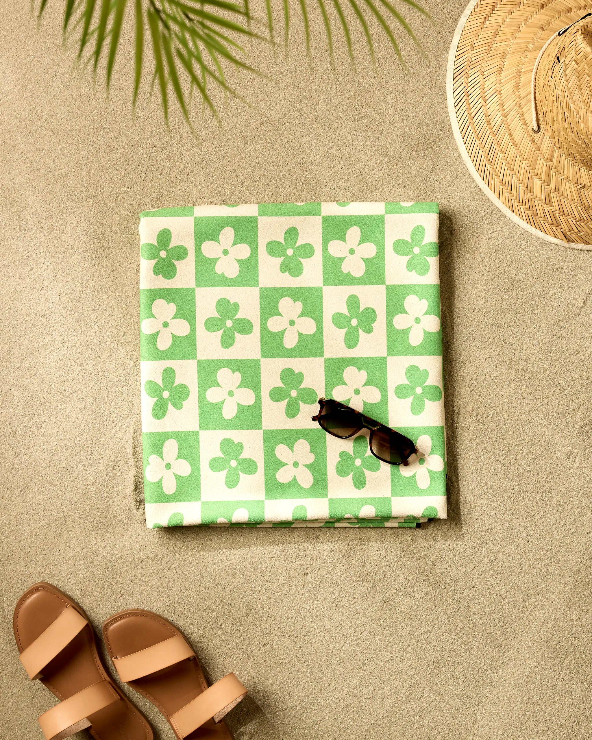 A pair of brown sandals, a woven straw hat, and sunglasses sit on sand next to a folded GEOMETRY Spearmint Green Flowers Micro-loop Beach Blanket; palm leaf shadows complete the scene.