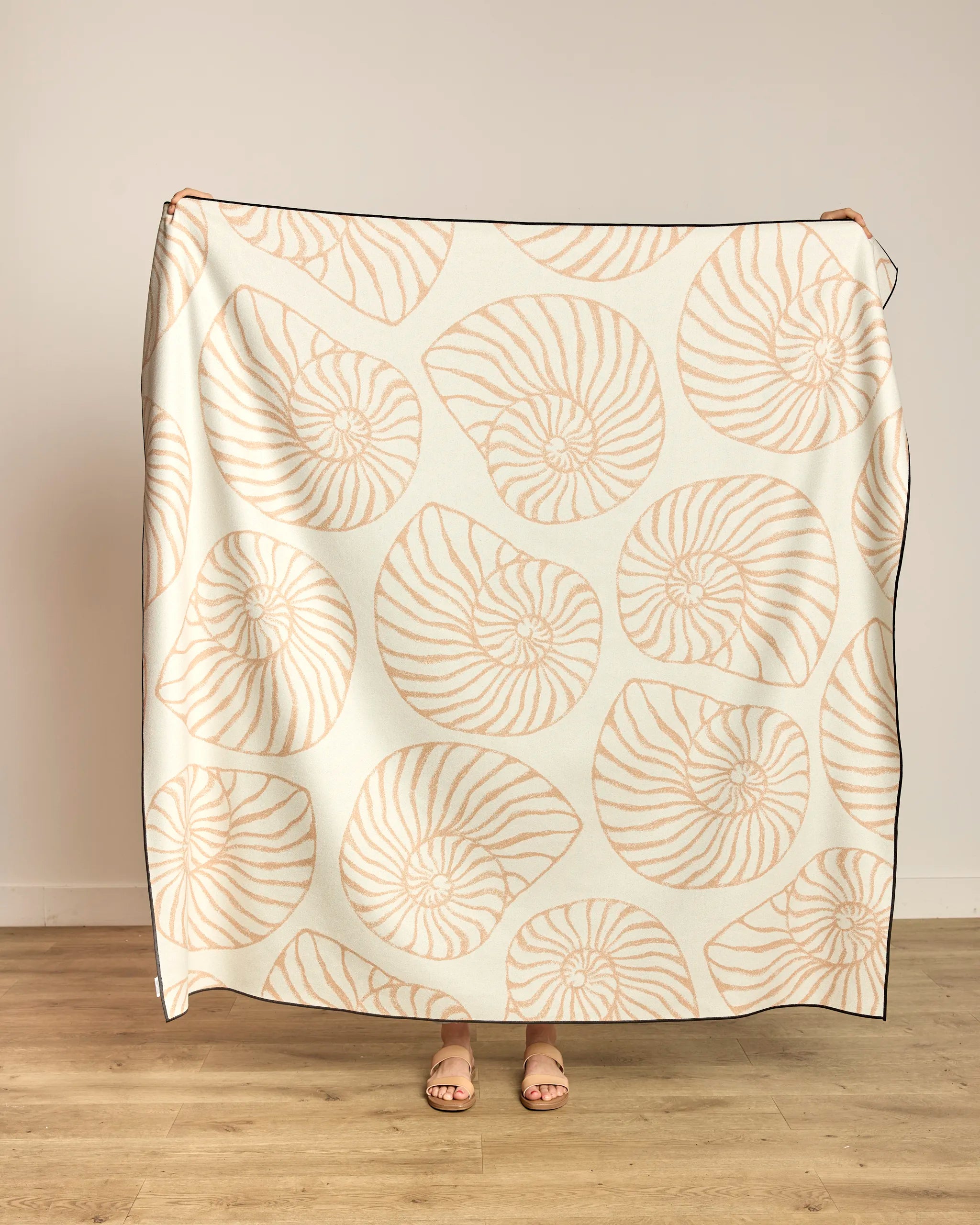 A person holds up the Spira beach blanket by Geometry, featuring a quick-drying cream fabric with a light tan nautilus shell pattern; only their hands and sandal-clad feet show against a plain wall and wooden floor.