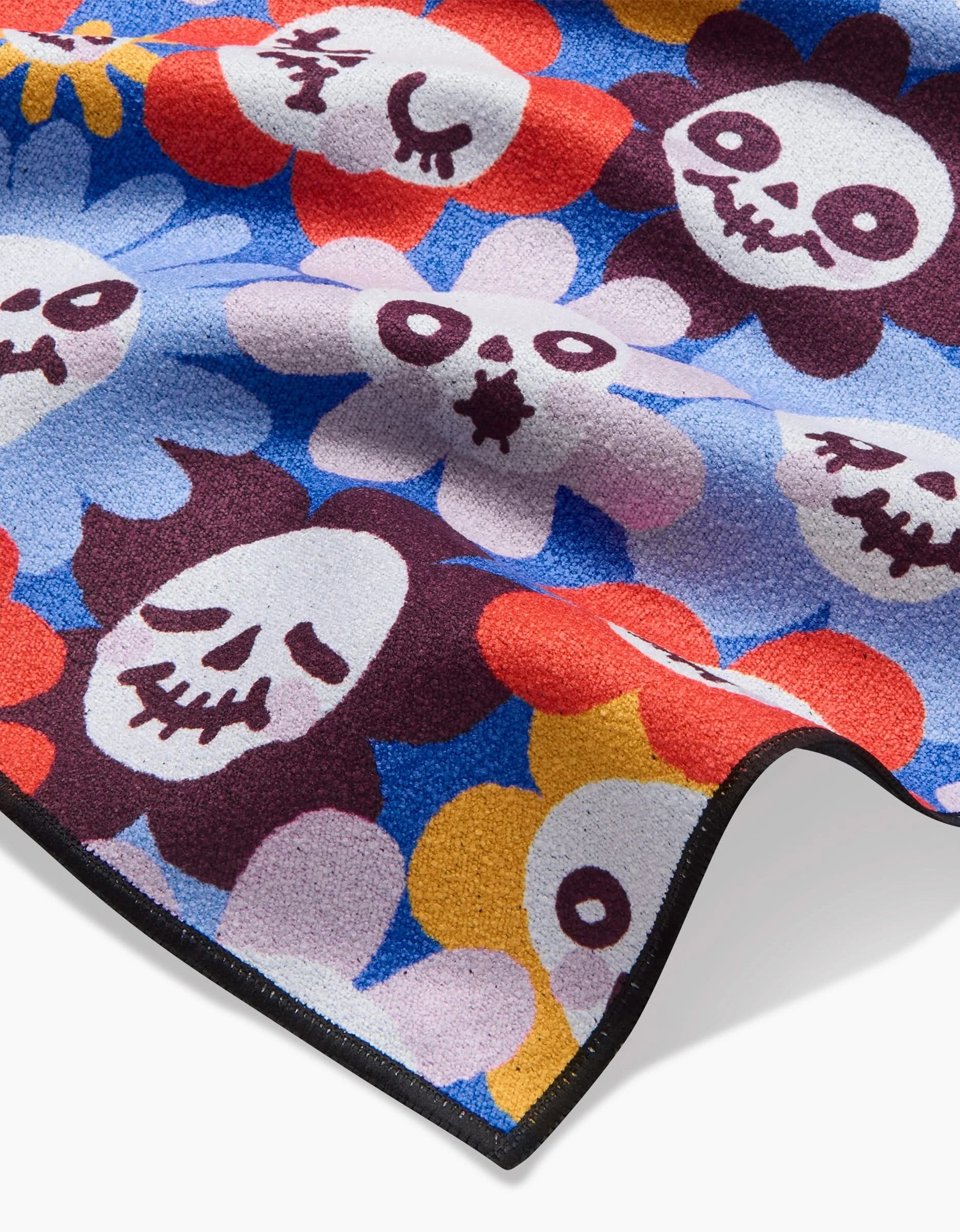 The So Lazo Spooky Bouquet beach blanket displays cartoon skull faces with flower petals in red, yellow, purple, and blue on a blue background. Made from quick-drying recycled fabric, it’s fun and eco-friendly with playful expressions and a visible edge.