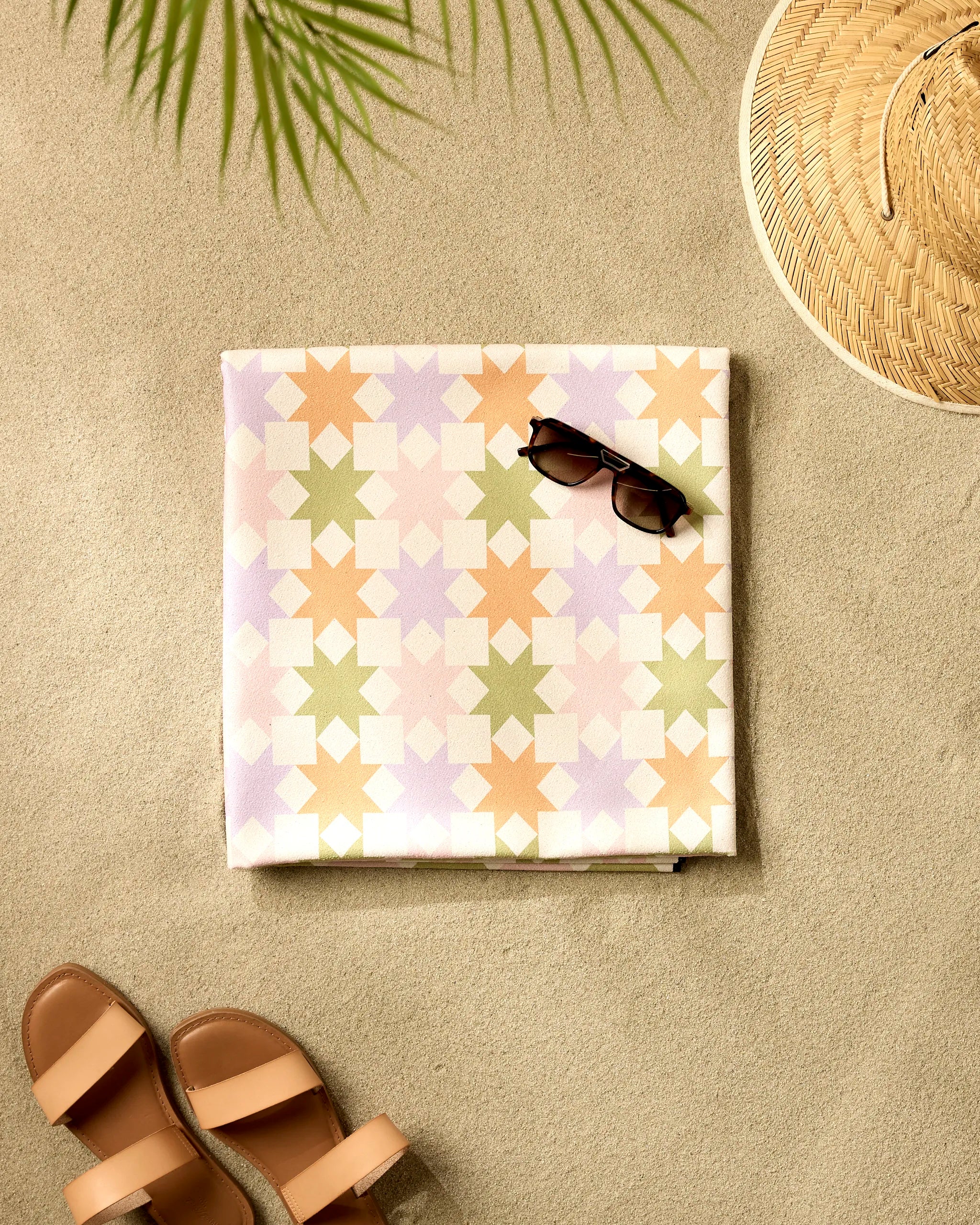 A GEOMETRY Spring Quilt with a pastel pattern is folded on the sand, topped with black sunglasses. Brown sandals, a straw hat, and a palm leaf shadow complete this inviting beach scene.