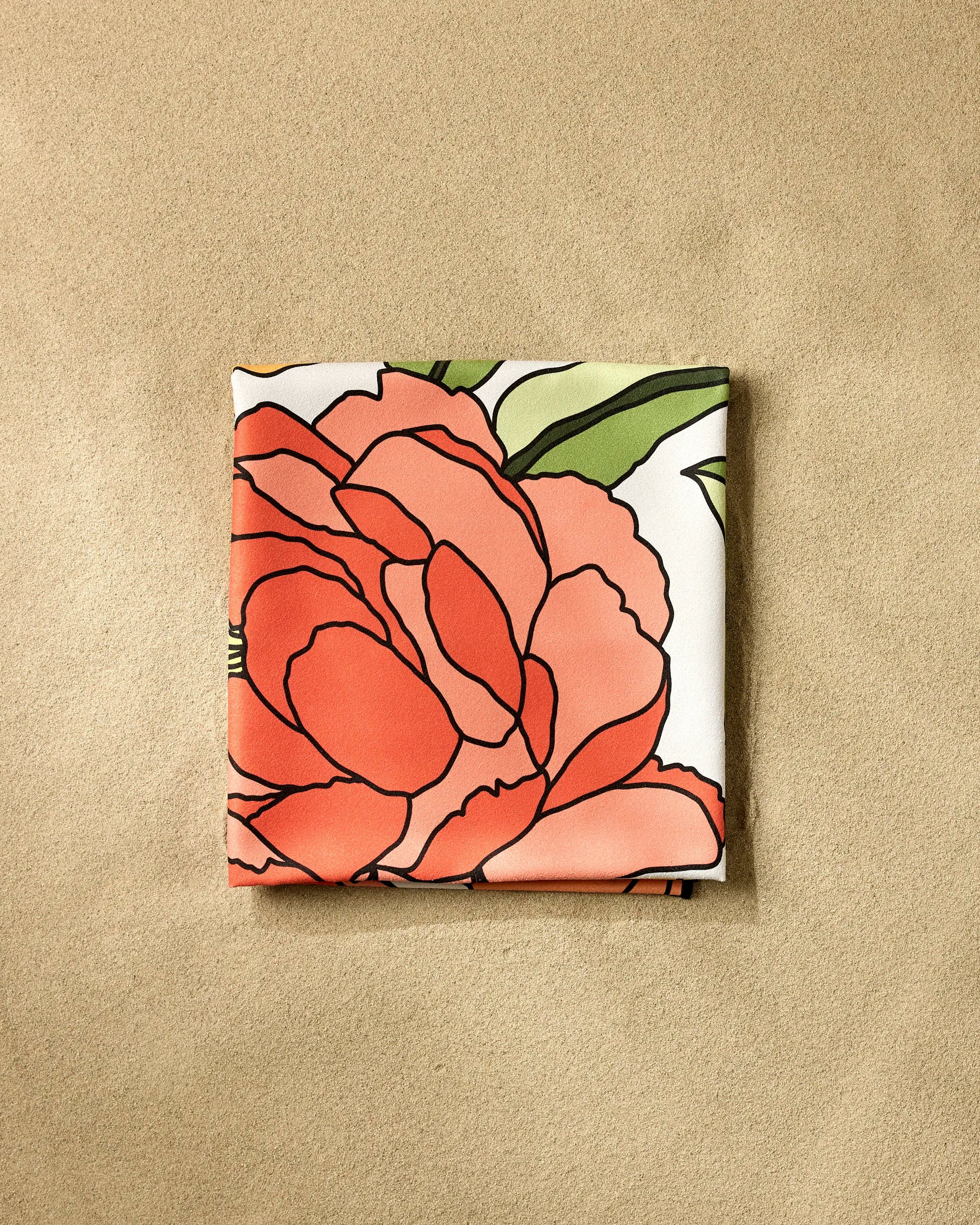 A neatly folded GEOMETRY "Summer Flowers" square scarf, featuring a bold red and coral floral print with green leaves, rests on a light beige textured surface, echoing the vibrant patterns of a Geometry Beach Blanket.