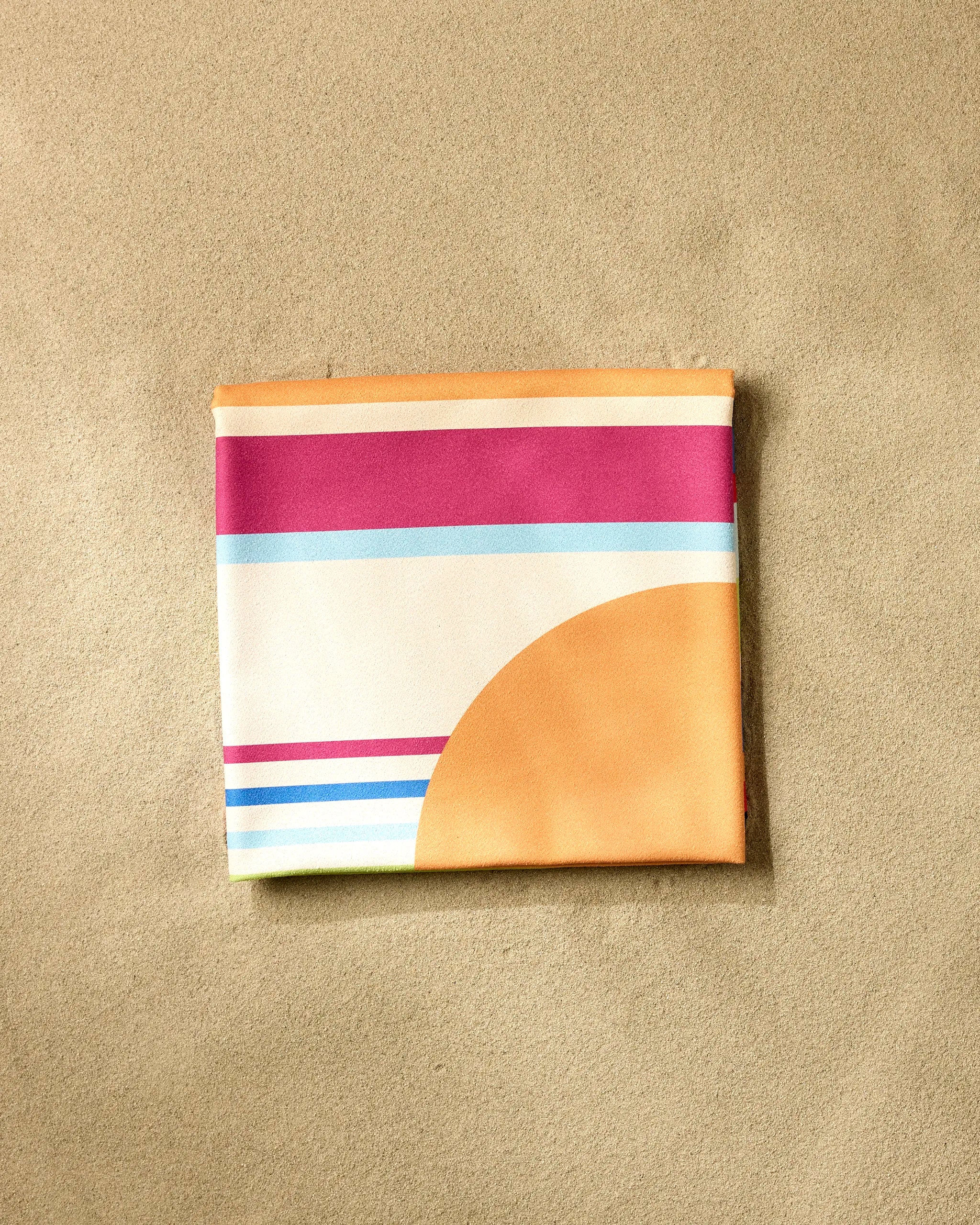The Sun And Stripes beach blanket by GEOMETRY, featuring vibrant orange, pink, teal, and blue geometric stripes and shapes, is shown folded on a sandy beige surface.