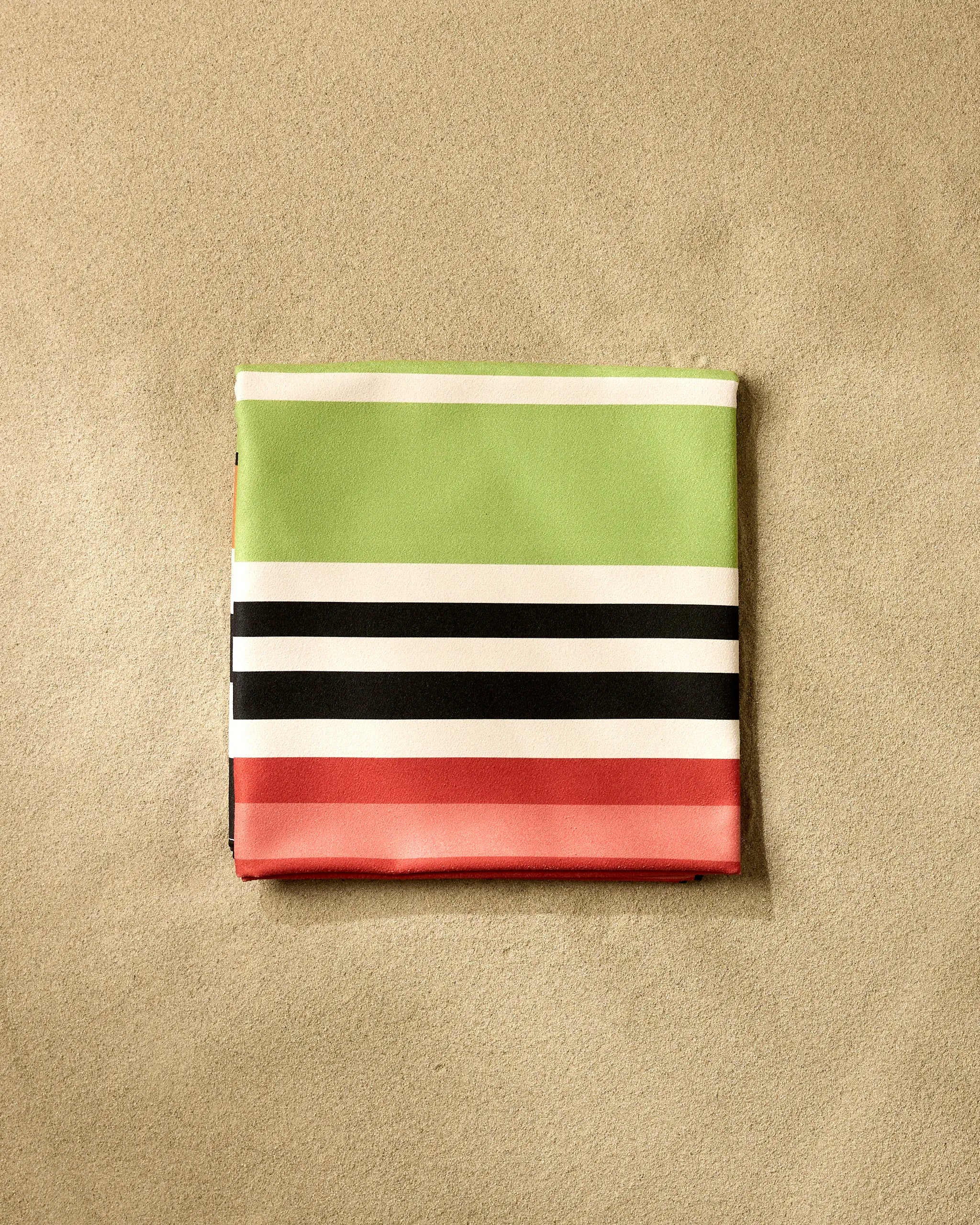 A neatly folded GEOMETRY Sunday Stripes Micro-loop Beach Blanket with green, white, black, and red horizontal stripes rests on sand, ready to provide quick-drying comfort for your next beach trip.