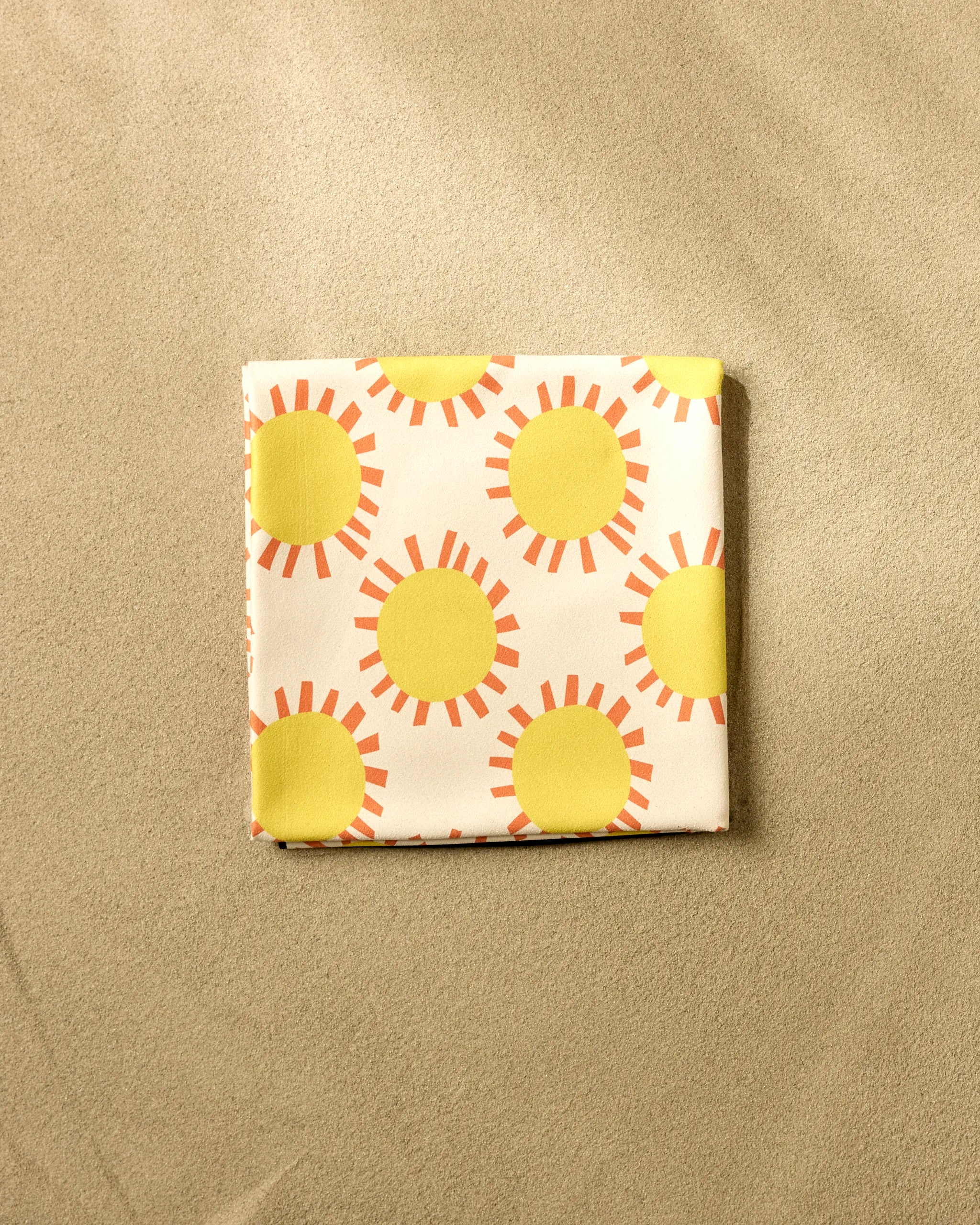 The Lizzie Clark Sunny Day Micro-loop Beach Blanket, quick-drying and comfortable, displays a bold yellow sun and orange ray pattern. Shown folded on a beige surface, it blends vibrant style with practical use.