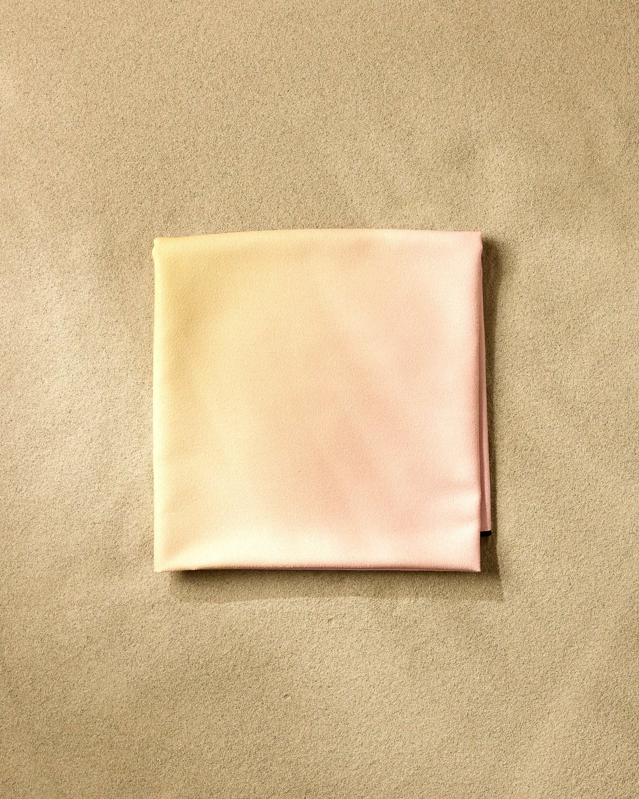 The GEOMETRY Sunset Gradient beach blanket, crafted from recycled materials, features soft yellow and pink hues and lies neatly folded on a textured beige surface—perfect for sustainable relaxation.