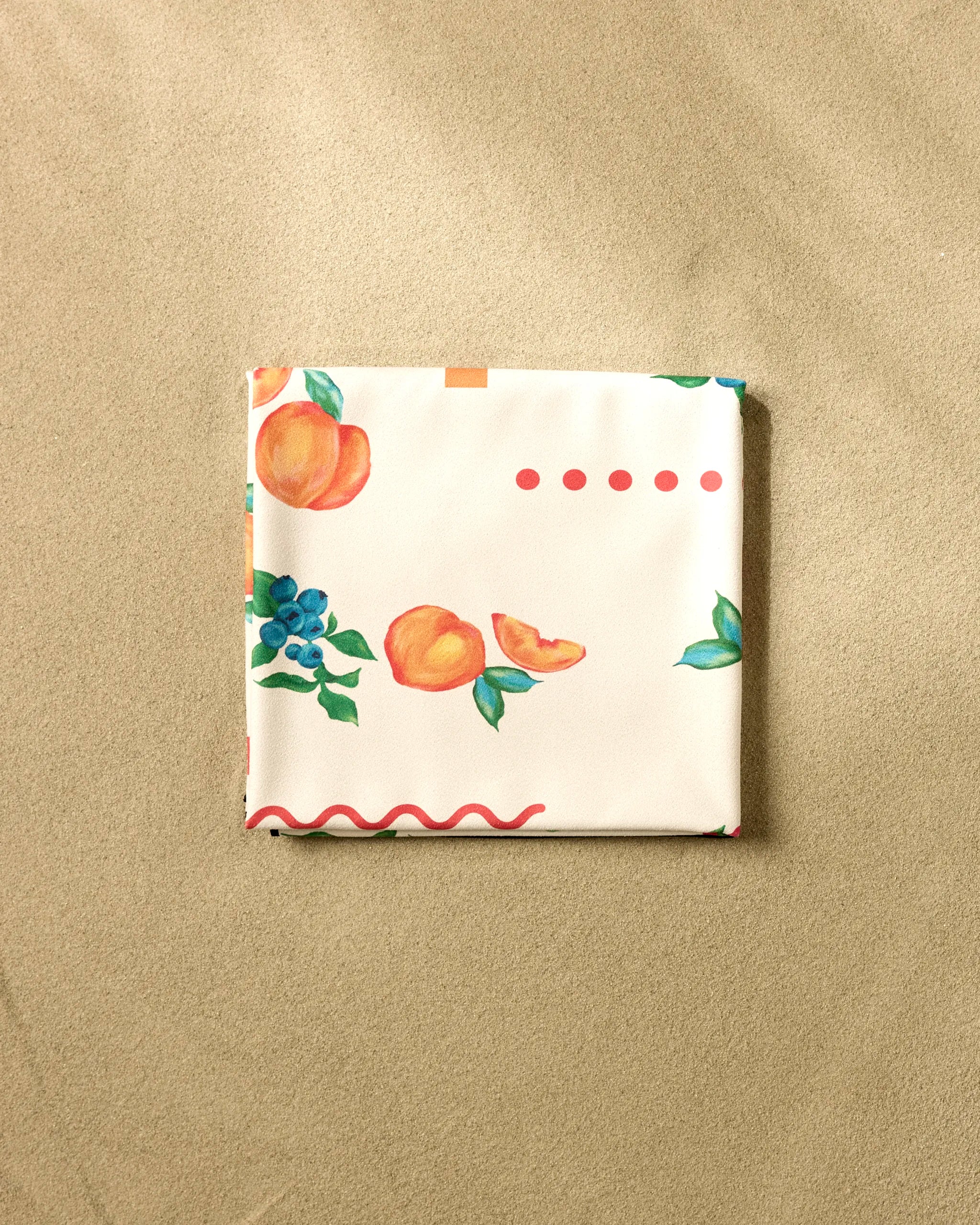 The "Sweet Pickings" beach blanket by Aubrey Fairchild, featuring colorful fruit illustrations and decorative pink and red lines, is shown folded on sand.