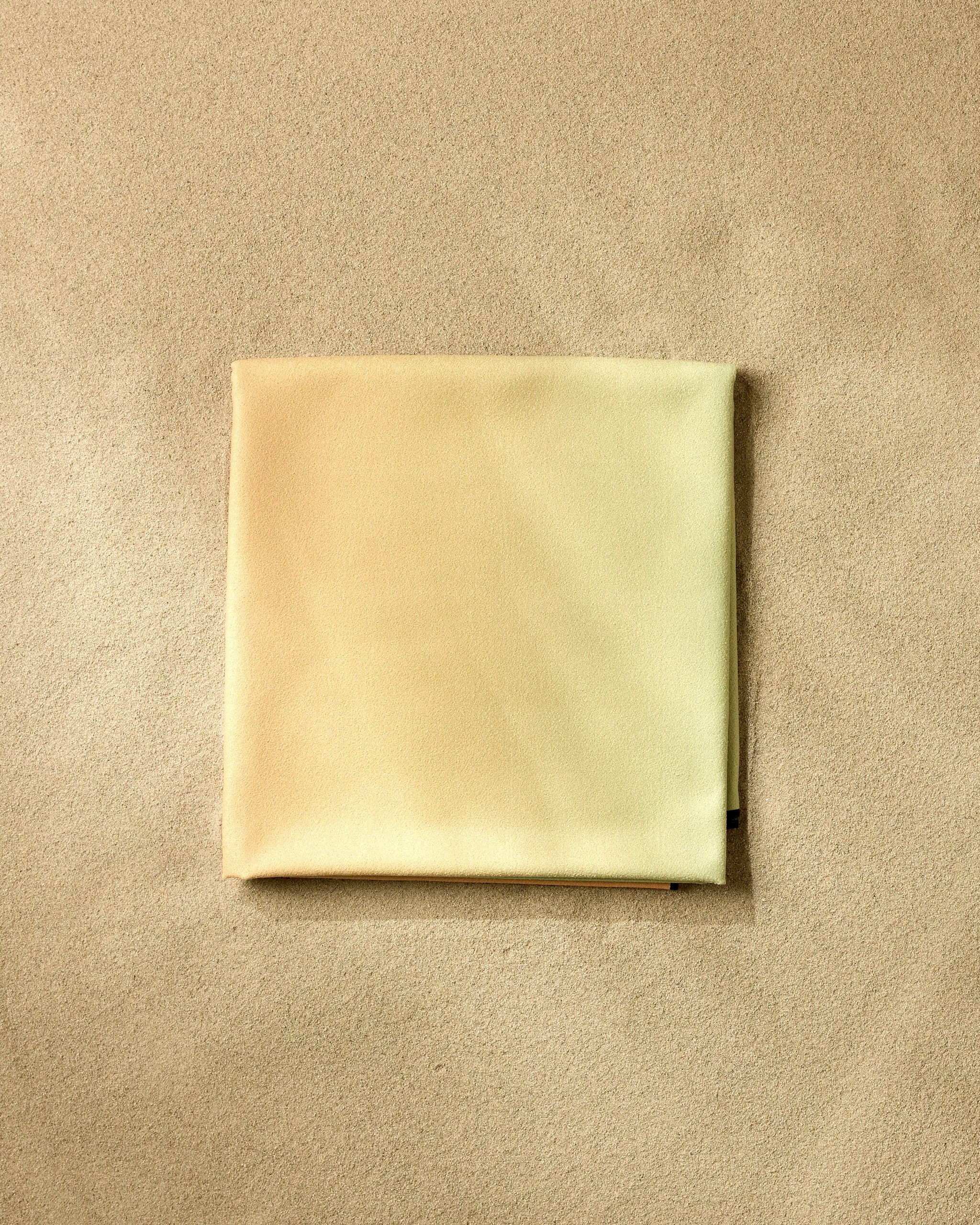 The Tangerine Dream beach blanket by GEOMETRY, crafted from recycled materials in light beige, is neatly folded on a sandy surface, blending subtly with its textured background.