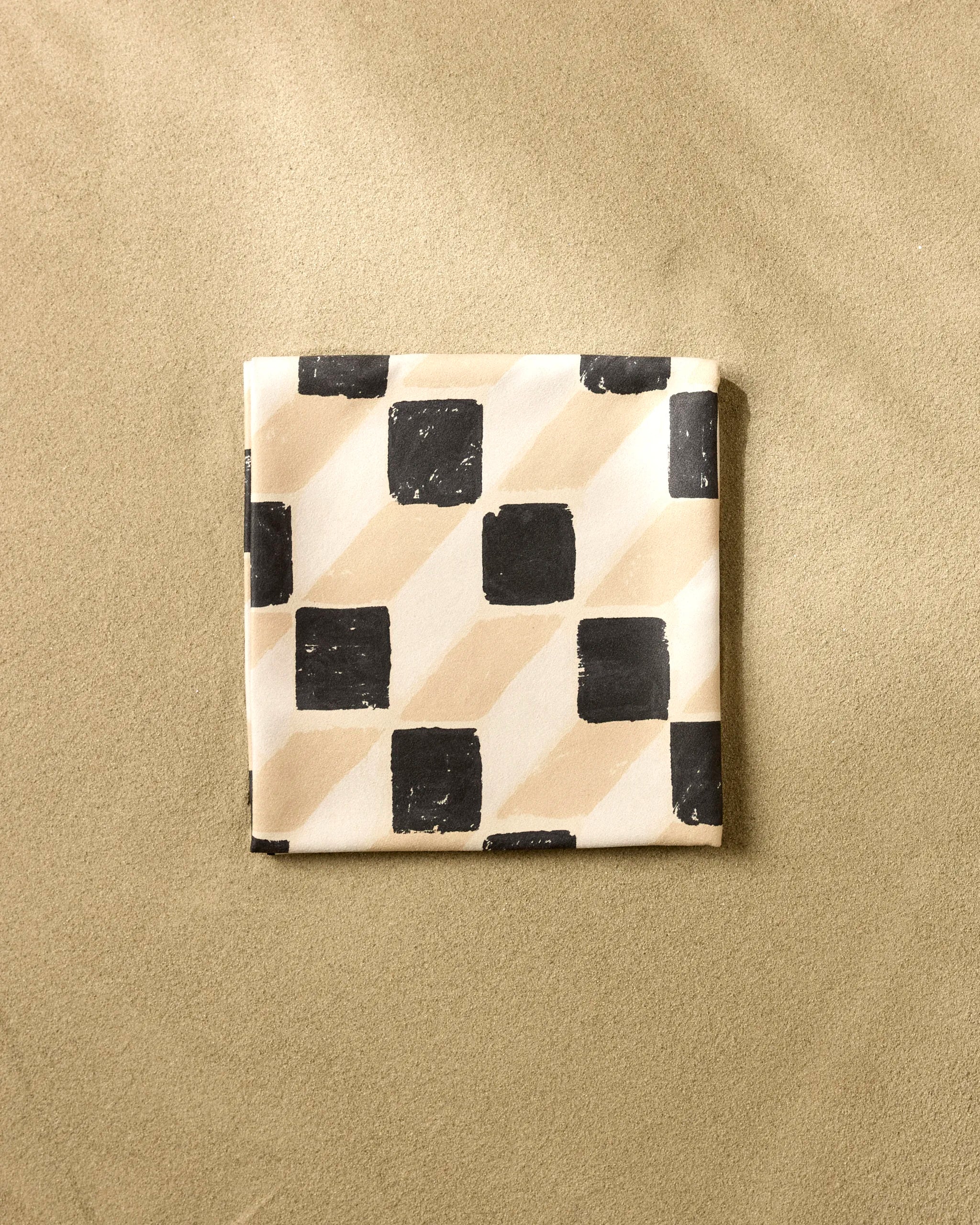 The Tessera napkin by Studio Jeannot features a black and beige checkered and diagonal striped pattern, crafted from recycled materials, and is shown neatly folded on a textured light brown surface.