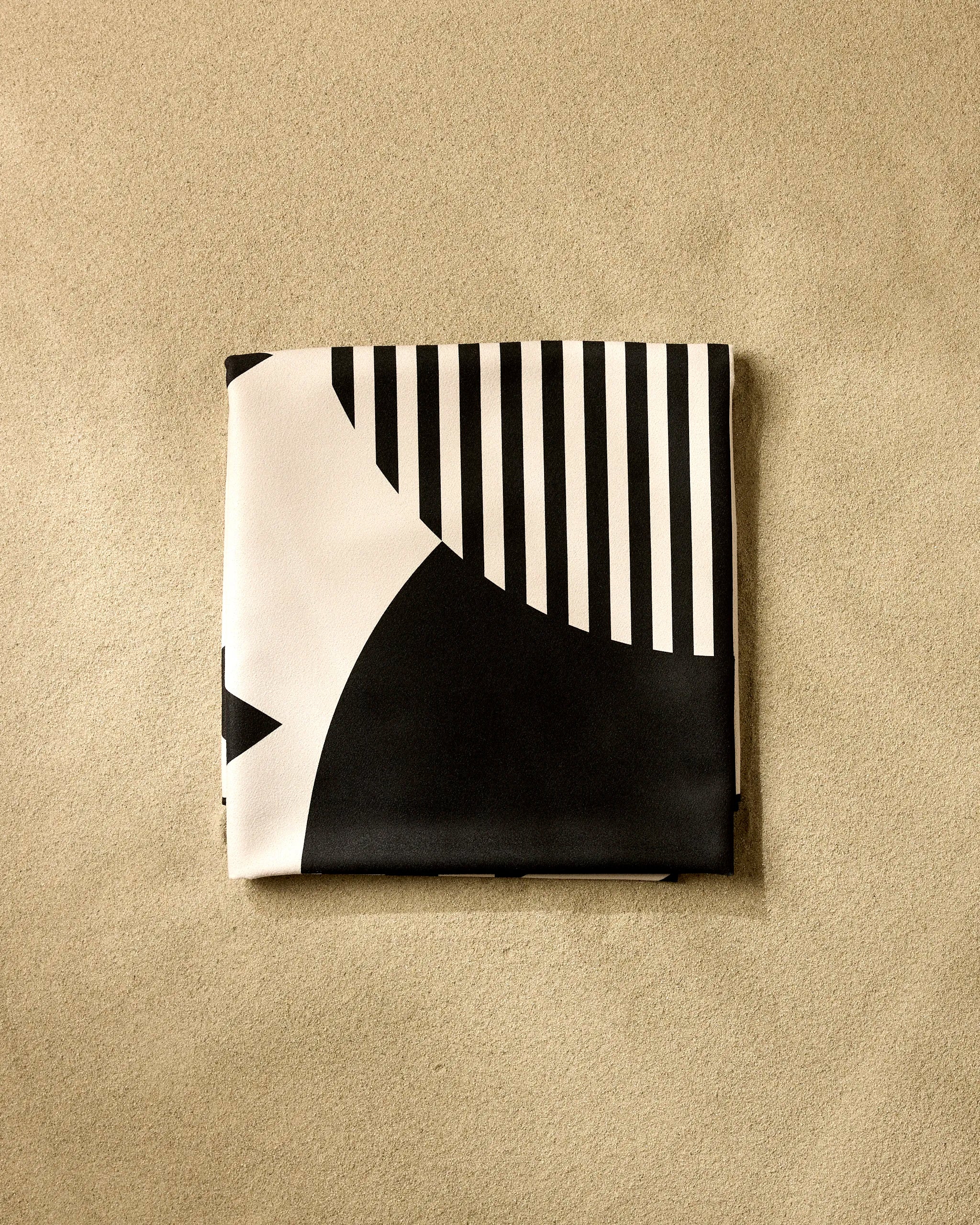 A neatly folded GEOMETRY The Outdoors beach blanket in black and white with bold geometric designs lies on textured beige—perfect as a quick-drying, stylish companion for days by the sea.