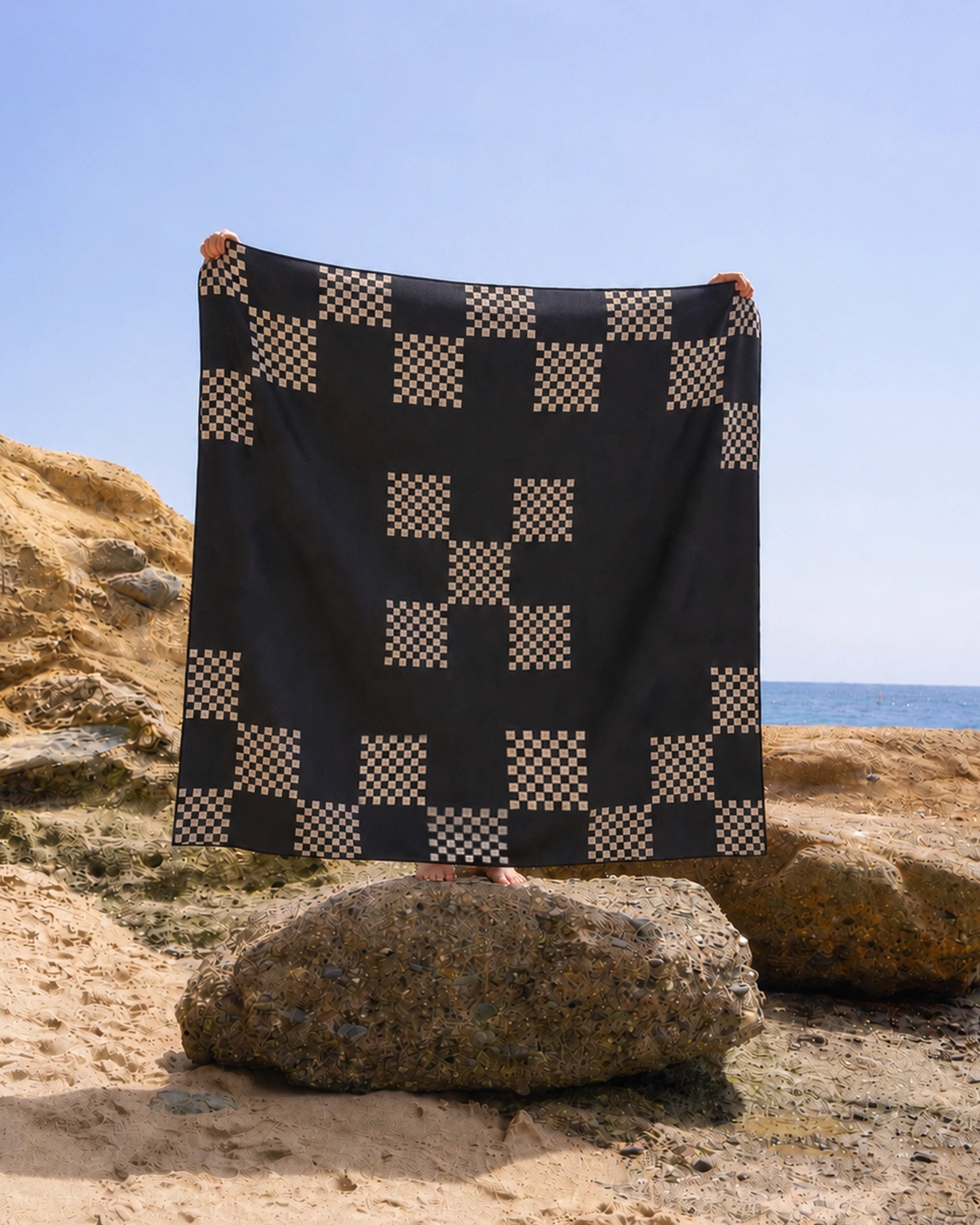A person stands on a large rock at the beach, holding up the Geometry Three in a Row Stone beach blanket, which features a black and beige geometric checker pattern. The clear blue sky and sea provide a serene backdrop.