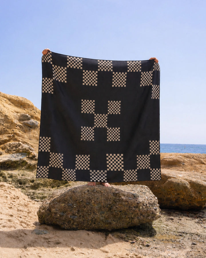 A person stands on a large rock at the beach, holding up the Geometry Three in a Row Stone beach blanket, which features a black and beige geometric checker pattern. The clear blue sky and sea provide a serene backdrop.