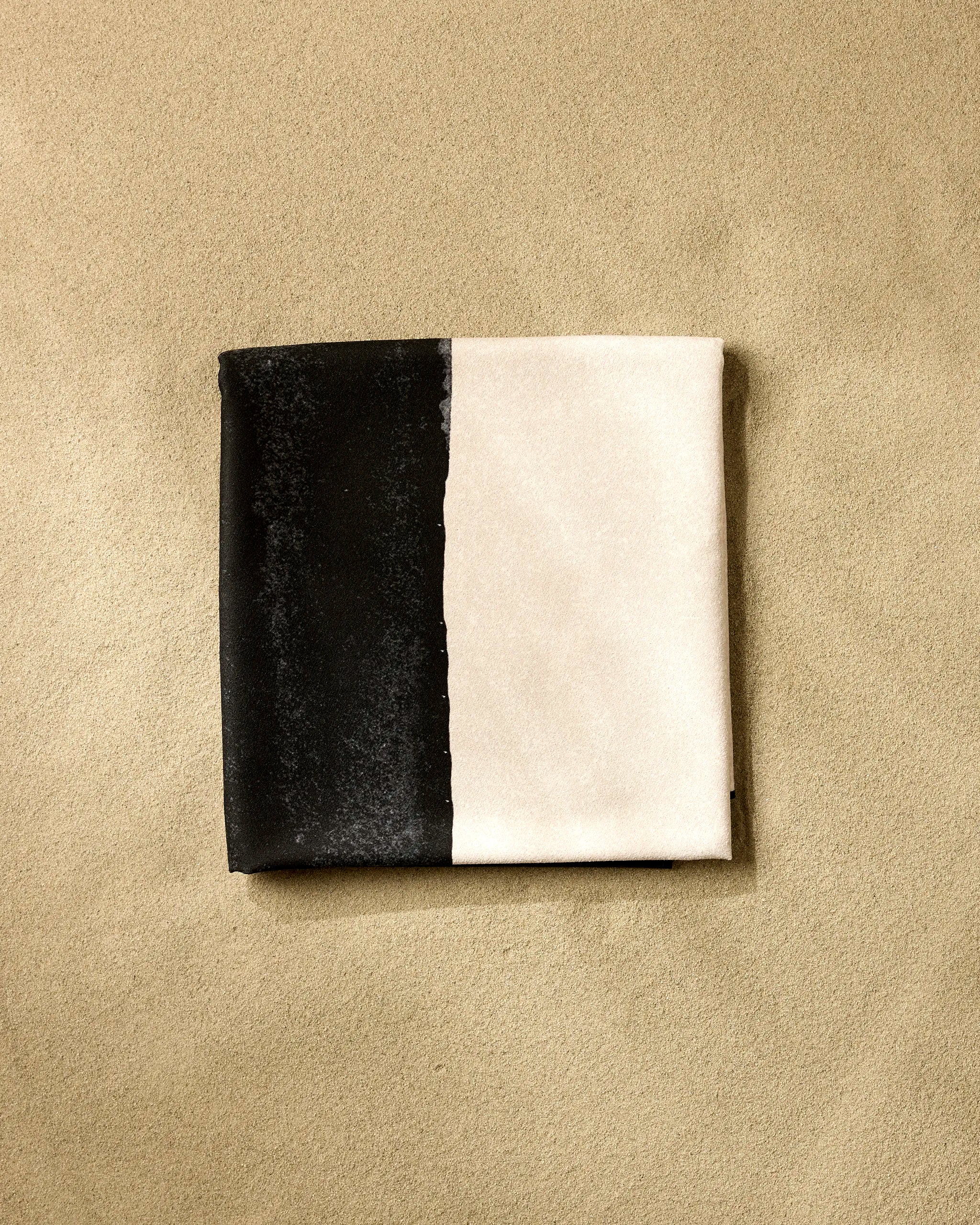 A neatly folded square napkin, inspired by the GEOMETRY Top Layer, features one half black and the other half white, resting on a sandy beige surface.