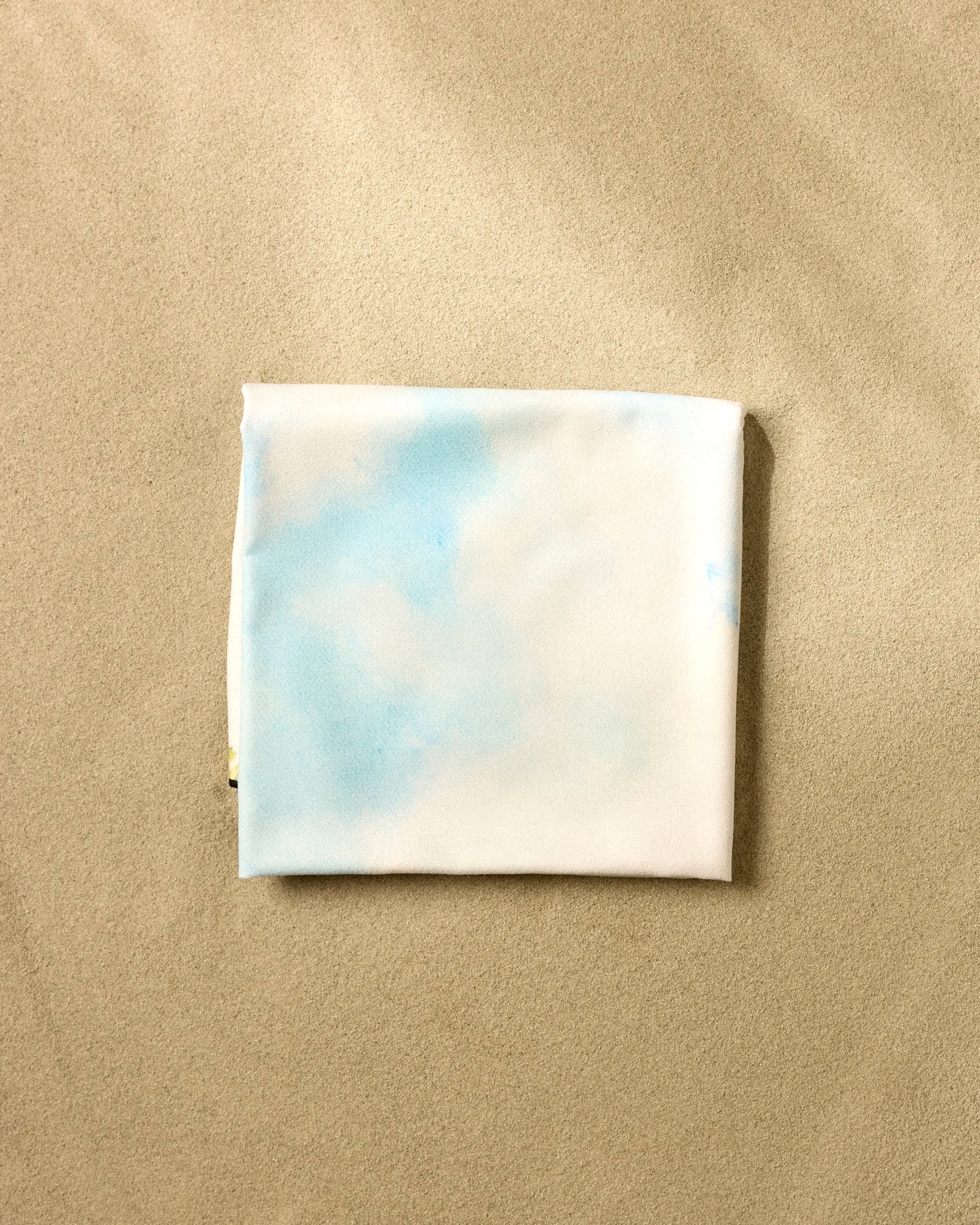 A neatly folded "Toward the Sun" beach blanket by Stefanie Bales, featuring light blue cloud-like patterns, rests on smooth beige sand.