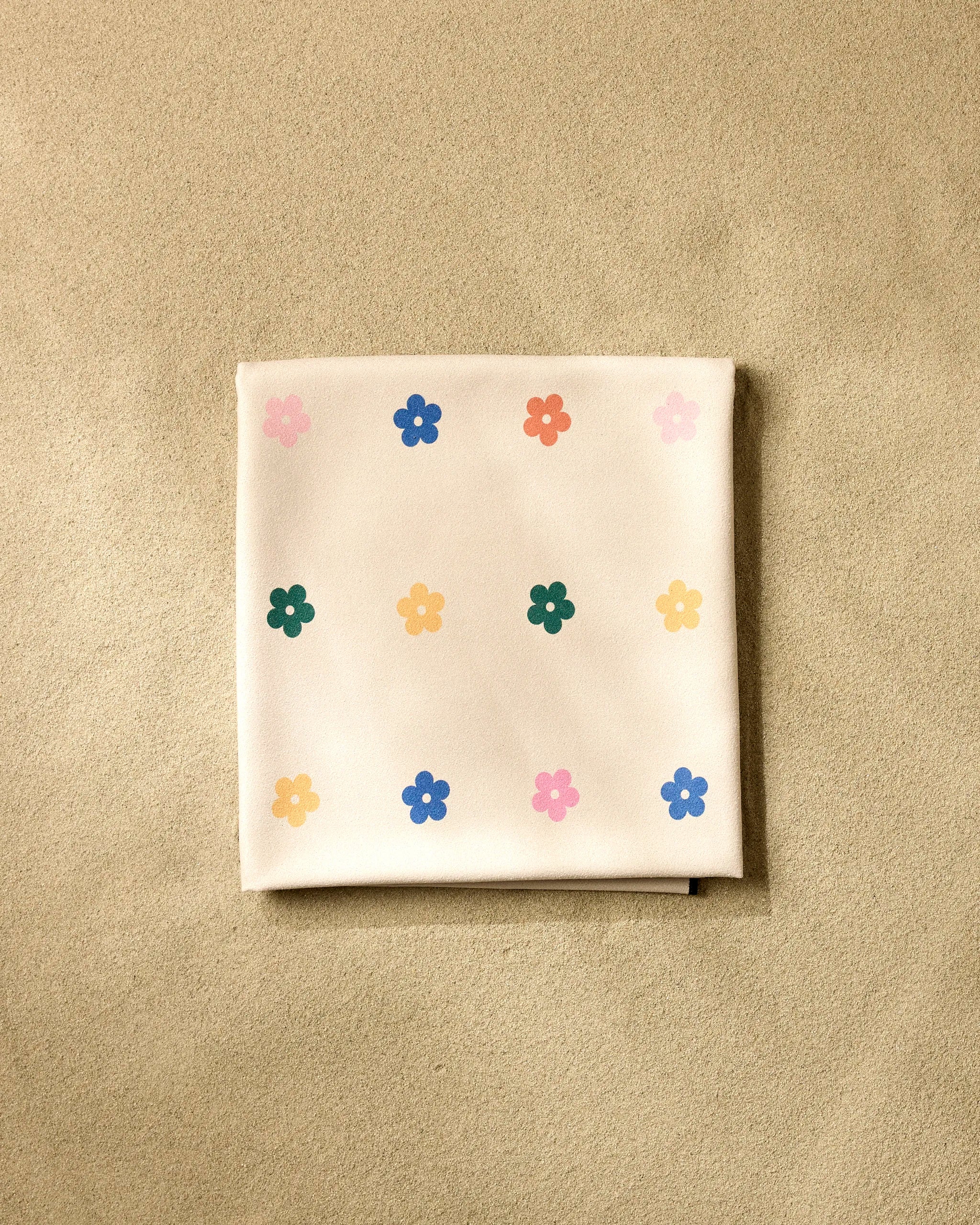 A folded GEOMETRY "Treat People with Kindness" beach blanket, crafted from recycled materials and featuring small colorful flower patterns, rests on a sandy beige surface.