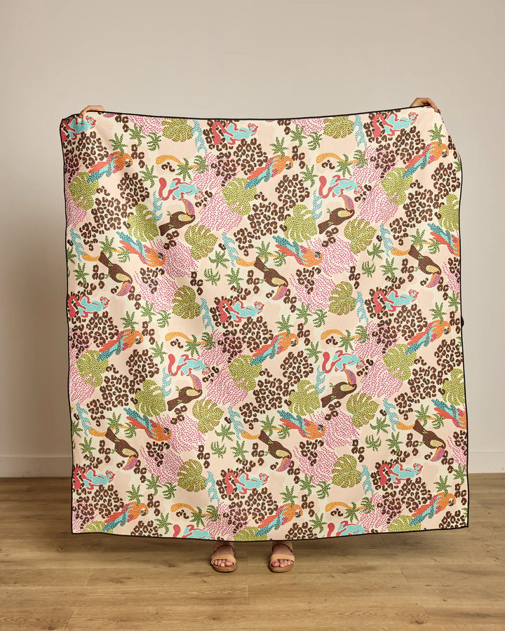 Wrapped in the Geometry Tropi Hotel beach blanket made from recycled materials, a person stands on a wooden floor against a beige wall, hidden behind its large, colorful pattern with only their feet showing.