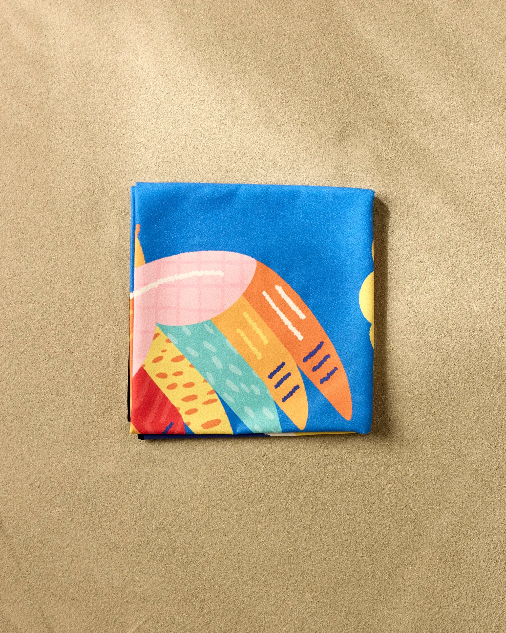 The Tropical Bird beach blanket by Camila Leão, featuring abstract patterns in blue, orange, yellow, red, and pink, lies folded on smooth light brown sand.