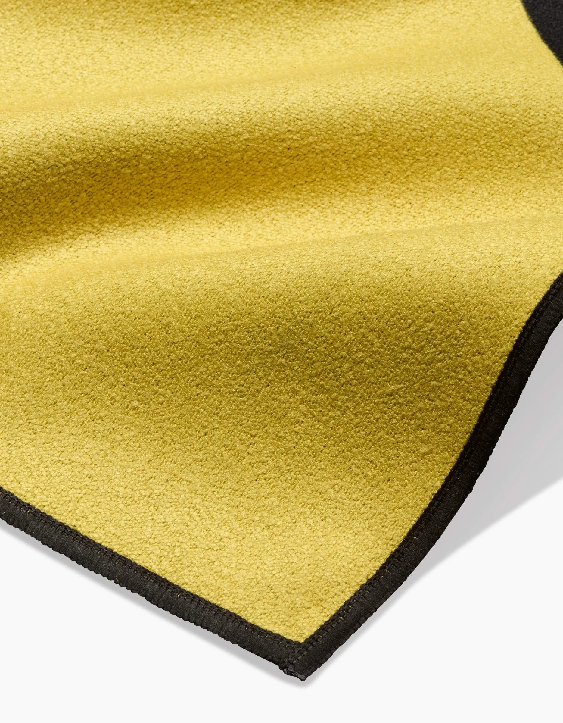 A close-up of the Camila Leão Tropical Bird yellow beach blanket, made from recycled materials with a soft, textured surface and black stitched border, slightly curved and folded on a white background.