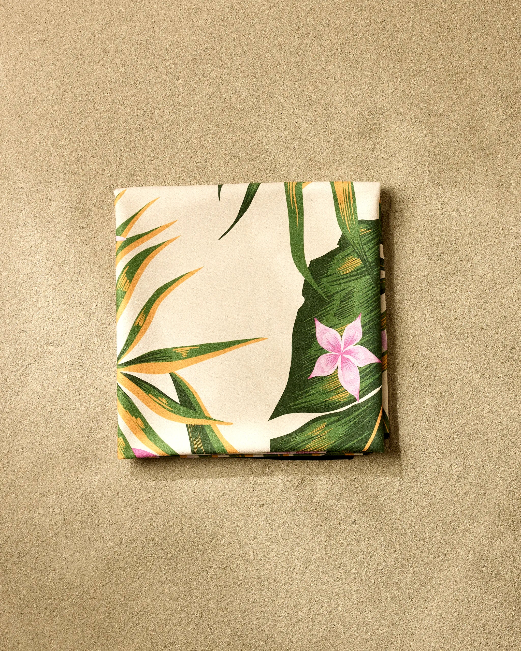 The GEOMETRY Tropical Jungle beach blanket, made from recycled materials, dries quickly and displays a tropical leaf and pink flower pattern on smooth light brown sand.