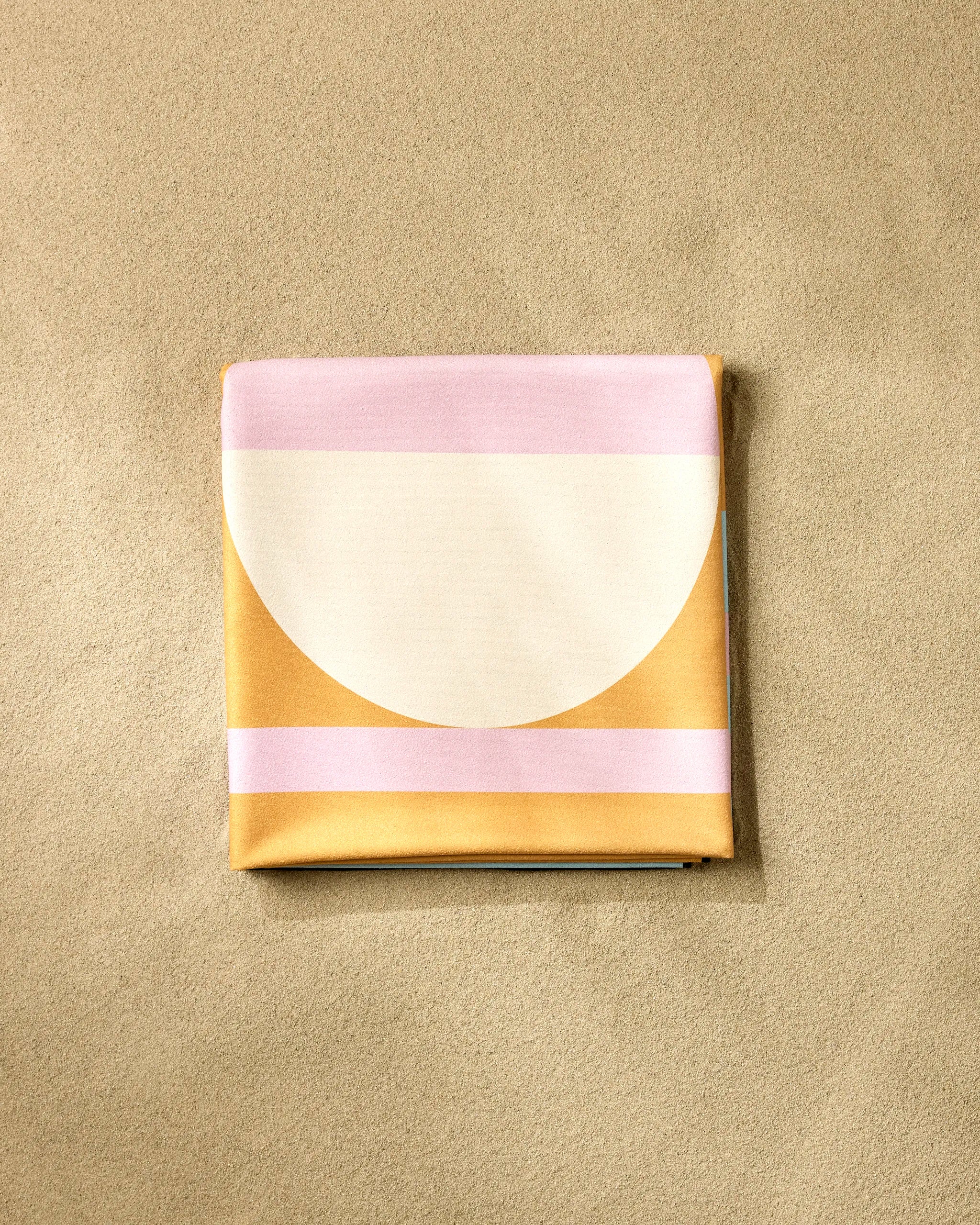 A neatly folded GEOMETRY Turmeric Pinks Beach Blanket, featuring geometric pink, white, and ochre shapes, sits on a beige textured surface.