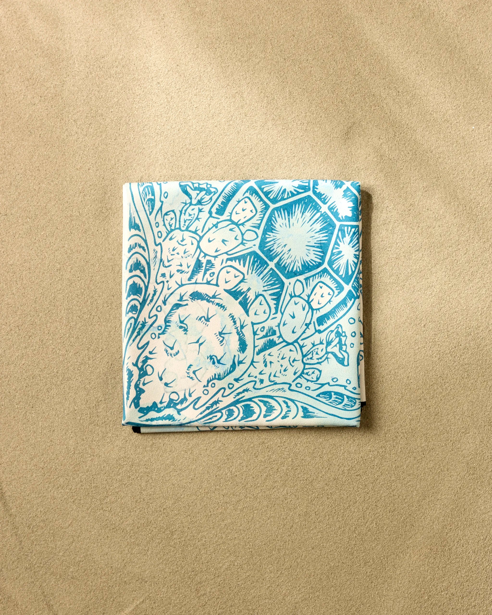 The Skye Walker "Turtle" beach blanket, featuring light blue and white abstract nature-inspired patterns and made from quick-drying recycled materials, rests on the sand.