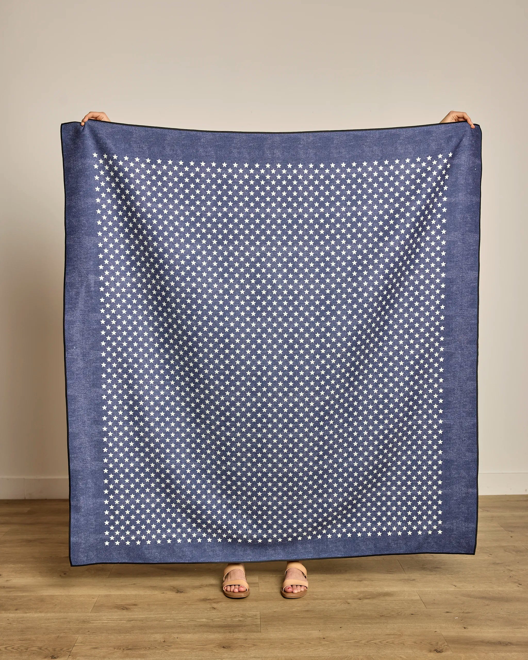 A person displays the Geometry Valor, a large blue quick-drying beach blanket with a white star pattern. Only their hands and feet are visible as they stand on a wooden floor against a plain light-colored wall.