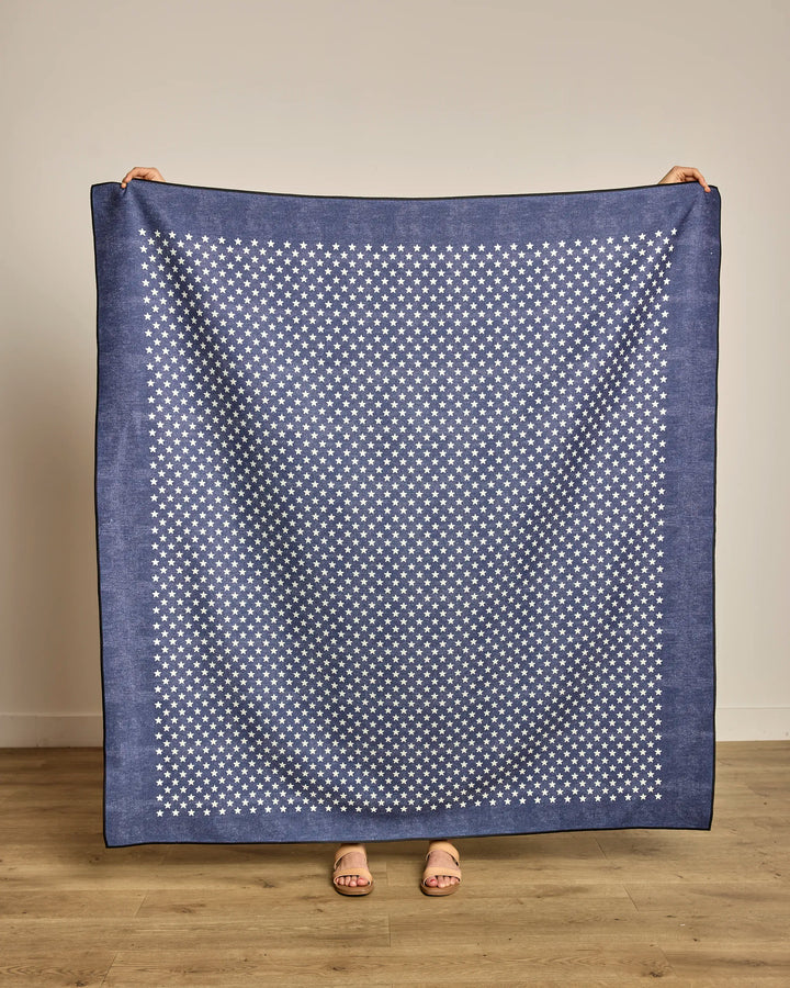 A person displays the Geometry Valor, a large blue quick-drying beach blanket with a white star pattern. Only their hands and feet are visible as they stand on a wooden floor against a plain light-colored wall.