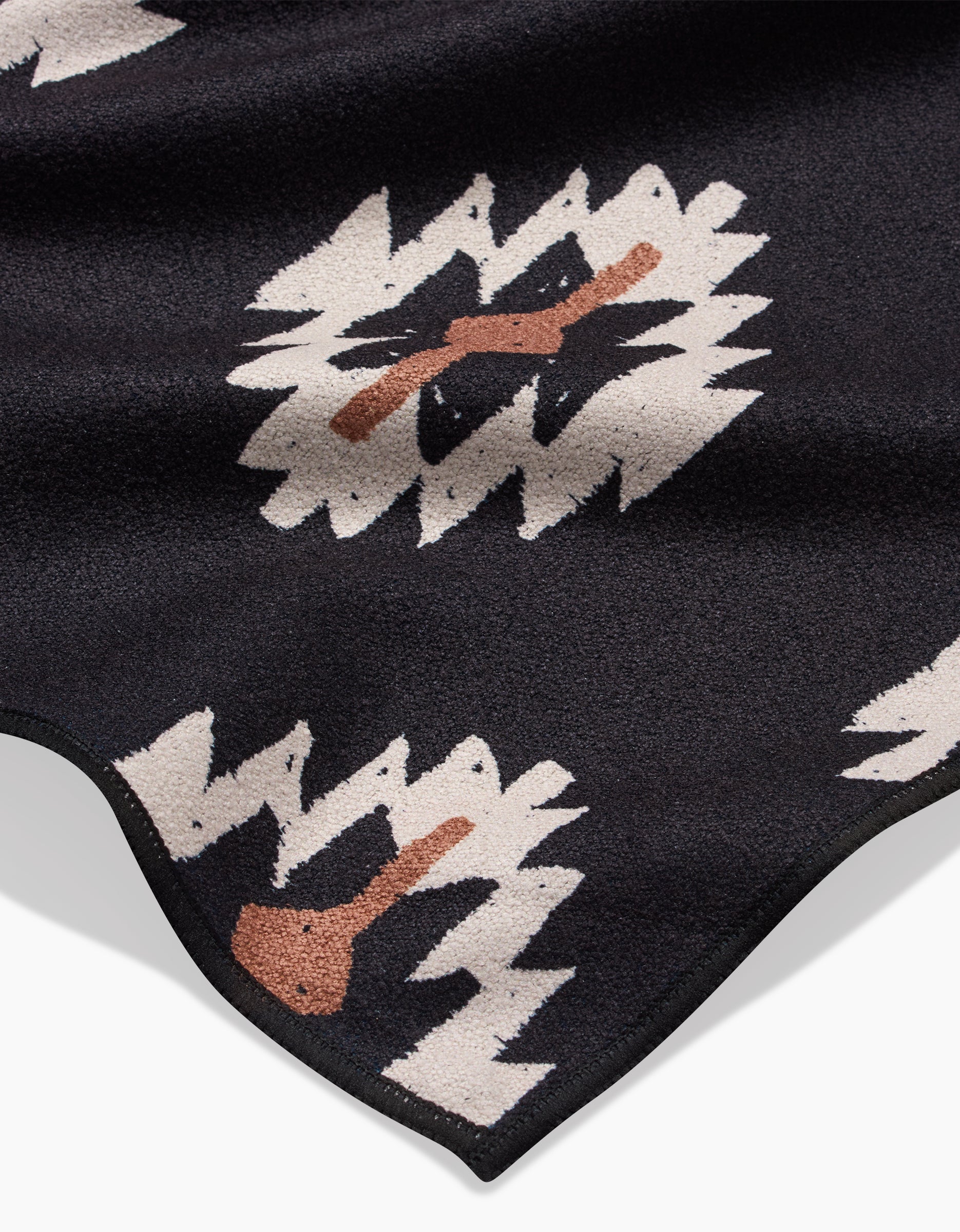 The Western Tile by Hufton Studio is a black fabric with a zigzag white pattern and brown abstract shapes, wavy edges, and a textured surface—this quick-drying beach blanket is crafted from recycled materials.