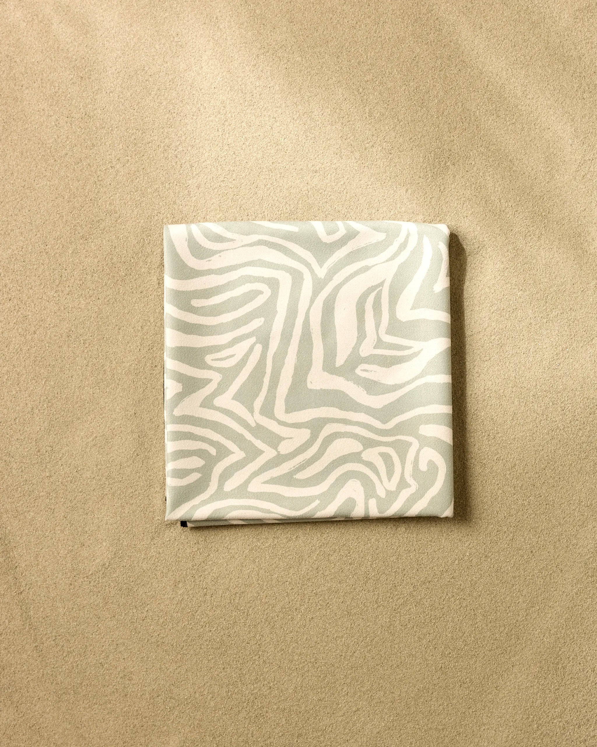 A neatly folded Hufton Studio Wild Marks Beach Blanket with a light gray and white abstract, zebra-like pattern lies on smooth, beige sand.