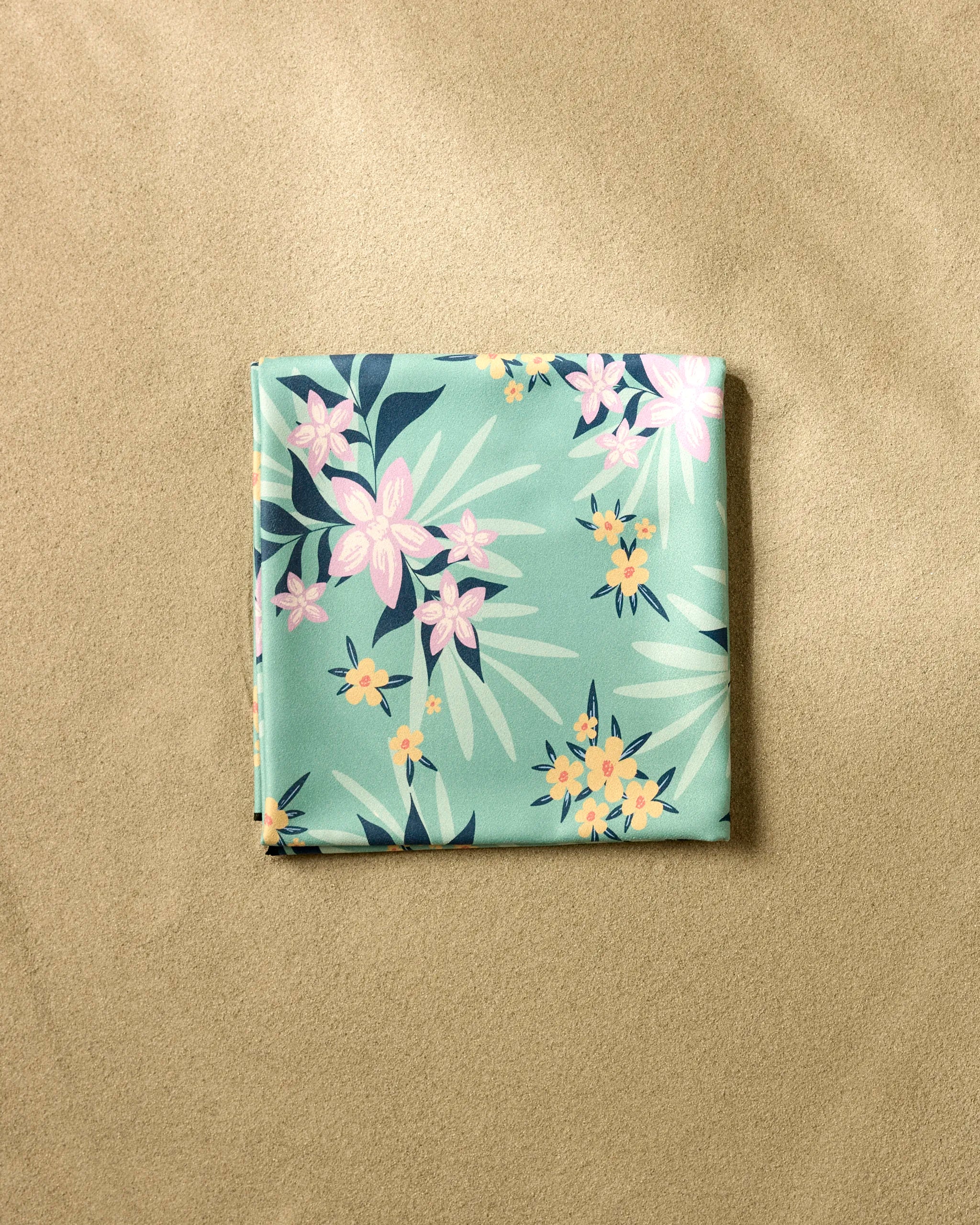 The Wilder beach blanket by Charlie Rowan Designs features a pastel-green color with pink and yellow floral patterns, made from recycled materials and neatly folded on smooth sand.