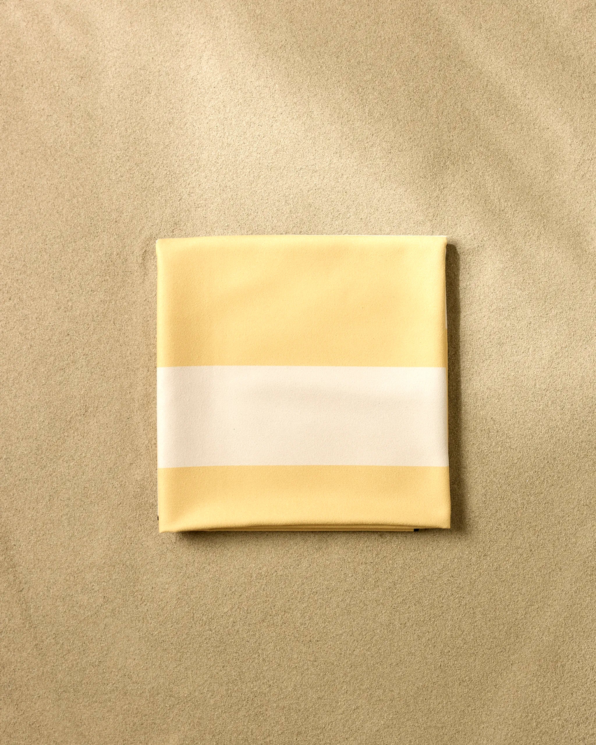 The GEOMETRY Yellow Block Stripes sand-repelling beach blanket, neatly folded, rests on smooth beige sand in a top-down view.