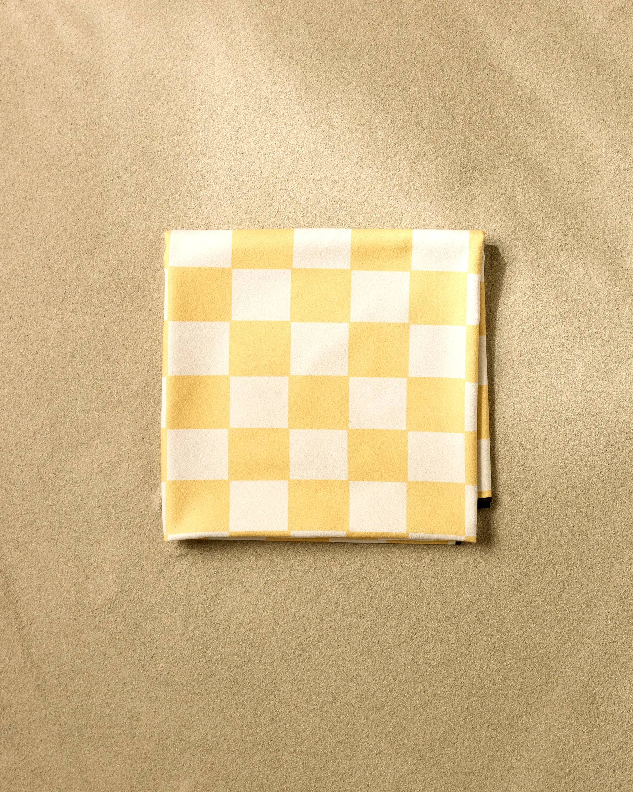 The GEOMETRY Yellow Checker Beach Blanket, featuring a folded yellow and white checkered design, rests on smooth sand—perfect for those seeking a stylish, sand-repelling blanket.