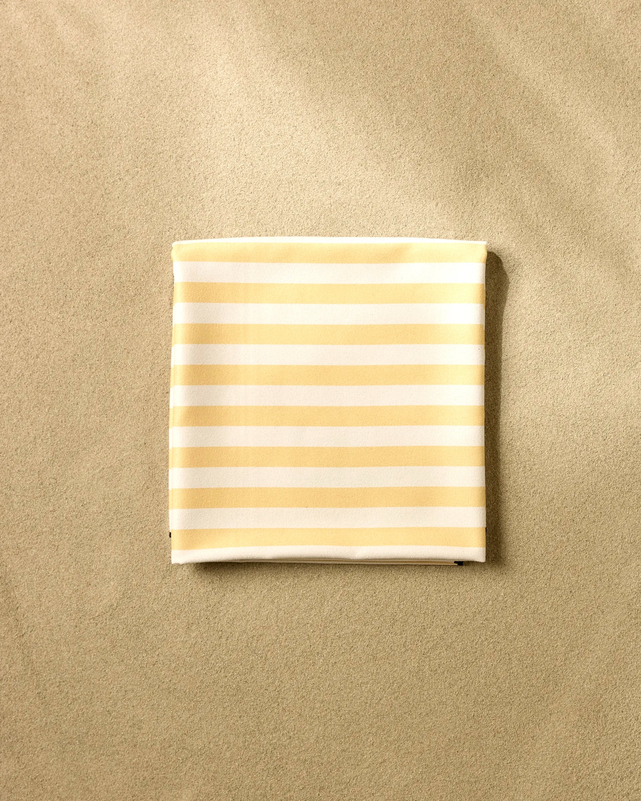 The GEOMETRY Yellow Stripes beach blanket, made from recycled materials, is neatly folded on smooth, light tan sand for a simple, minimal look.