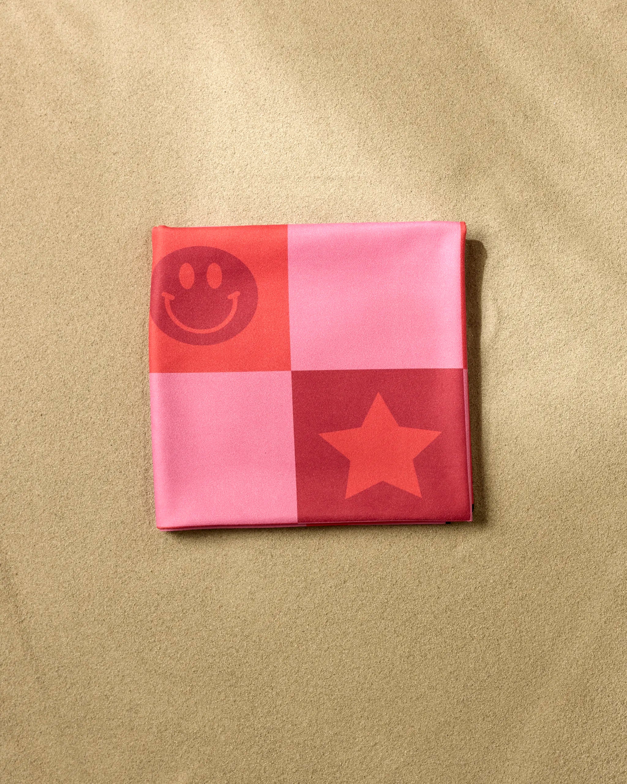 A folded pink and red "You're Doing Great" Micro-loop Beach Blanket by Quotes by Christie, featuring smiley face and star patterns, sits on light brown sand.
