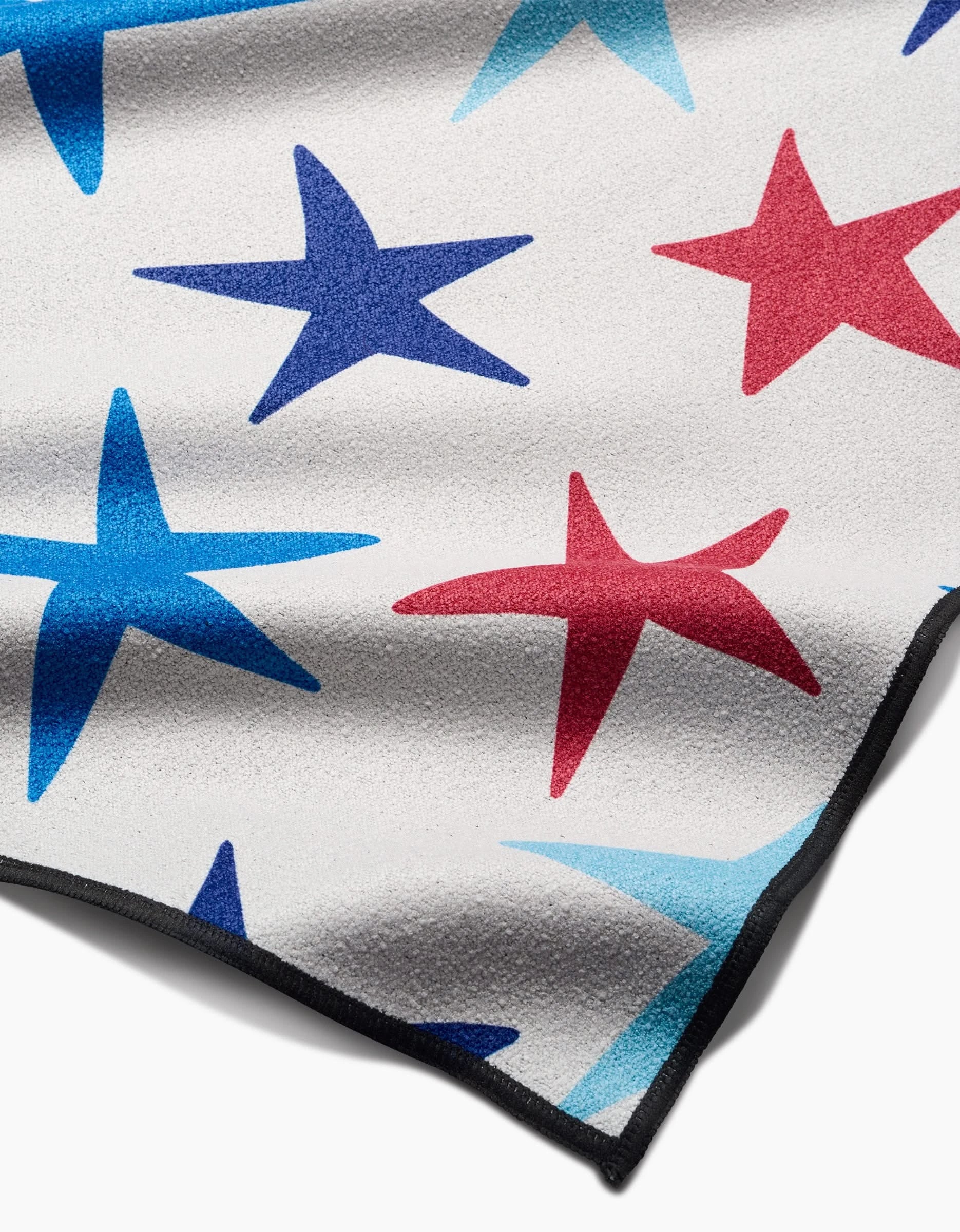 A close-up of Mindfulnices Seeing Stars Micro-loop Beach Blanket, featuring a white base with red, blue, and light blue stars, visible texture, black-trimmed edges, and crafted from recycled materials.