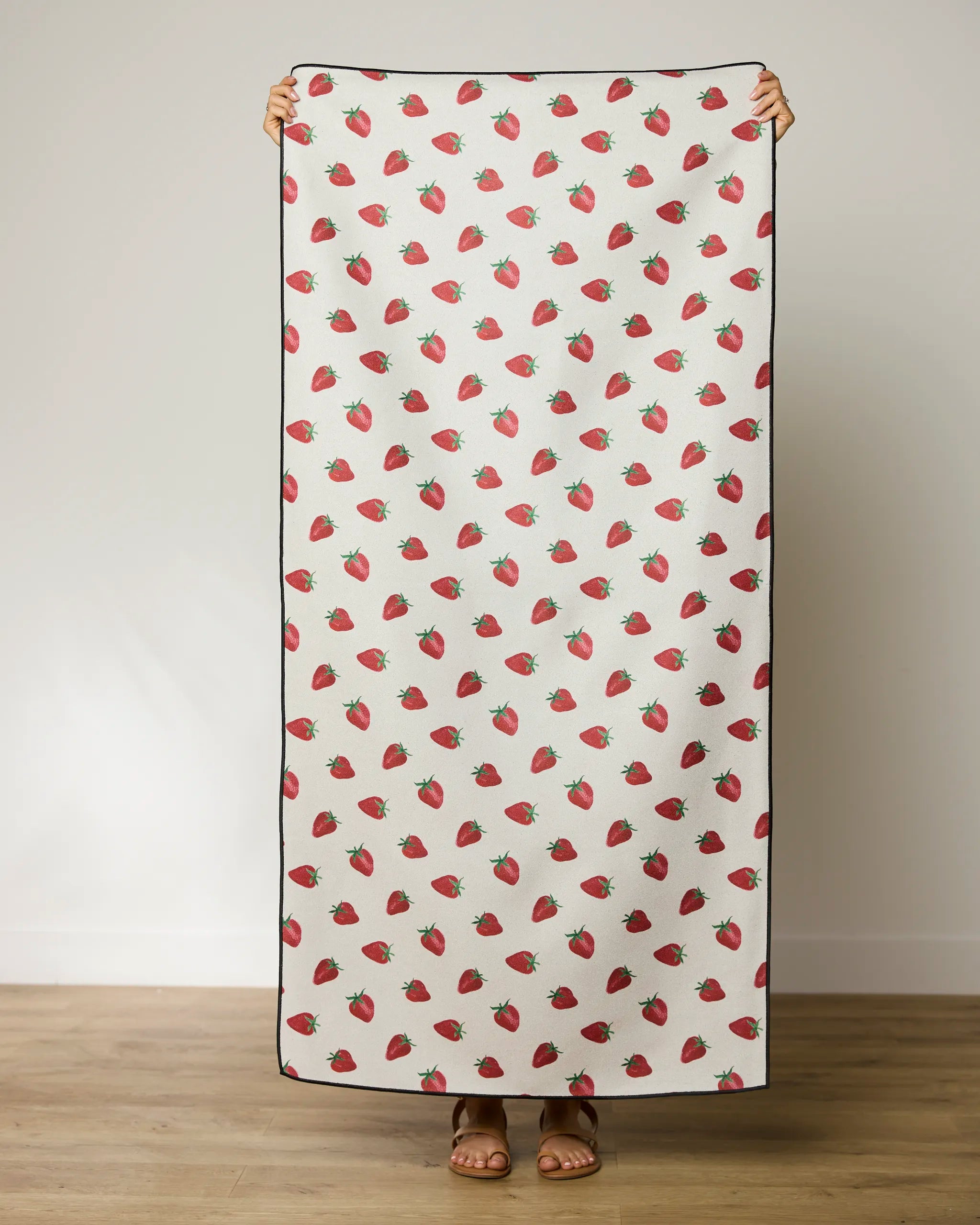 A person stands indoors holding Geometry’s Berry Patch quick-drying beach towel, featuring a white background with scattered red strawberries; only their hands, feet, and sandals are visible.