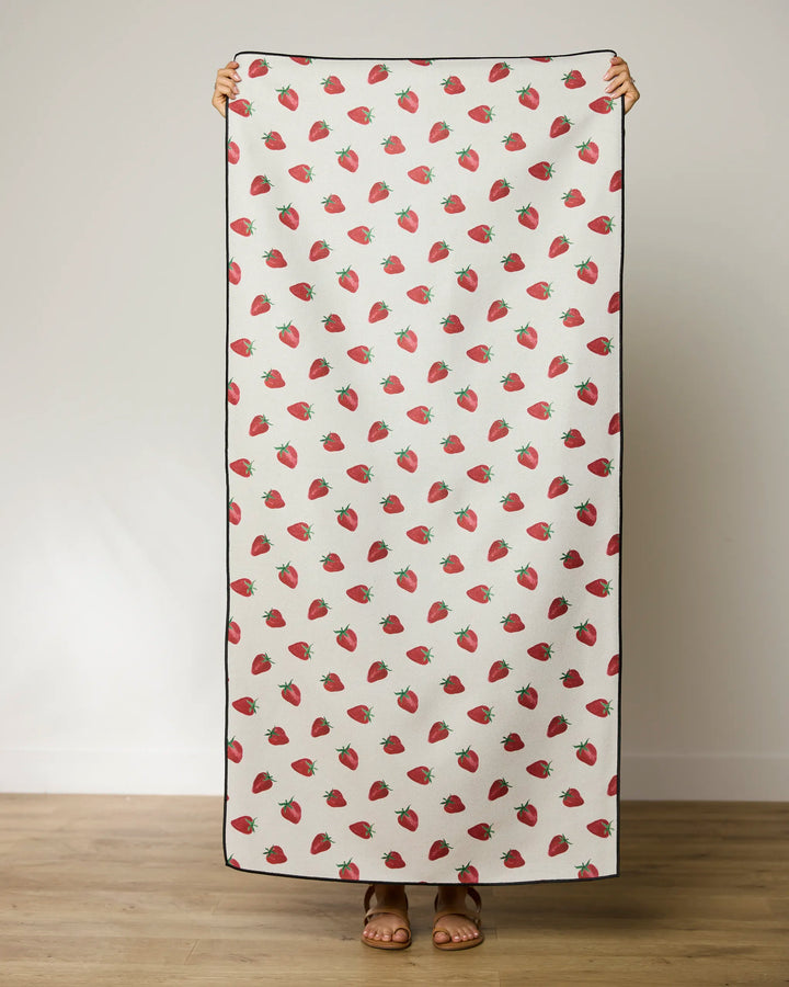 A person stands indoors holding Geometry’s Berry Patch quick-drying beach towel, featuring a white background with scattered red strawberries; only their hands, feet, and sandals are visible.
