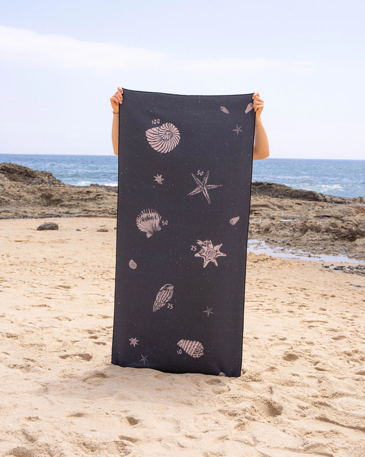 A person stands on a sandy beach holding a Geometry Bullseye Stone Micro-loop Beach Towel. Seashell and starfish designs decorate the towel. In the background, rocky shoreline and ocean waves appear beneath a partly cloudy sky.