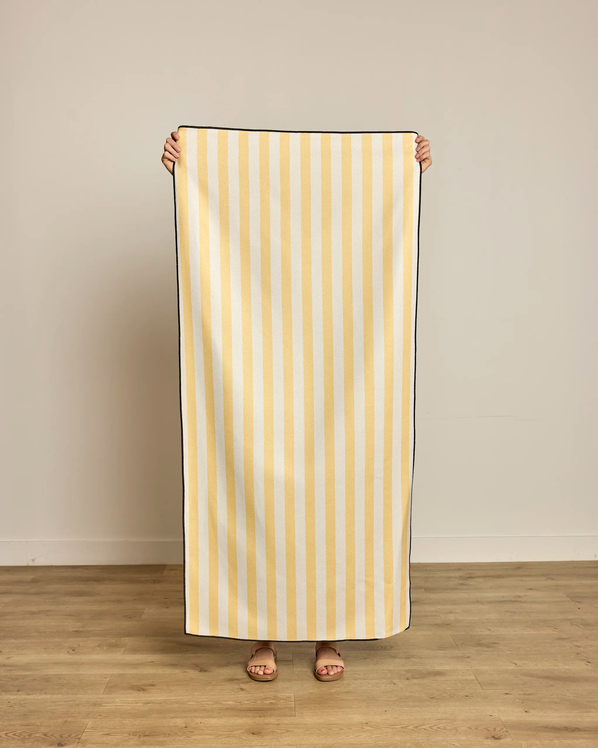 A person stands on a wooden floor holding up the Geometry Cabana Micro-loop Beach Towel, featuring bold vertical yellow and white stripes. The quick-drying towel pops against the plain, light-colored wall, obscuring their body except for their feet.
