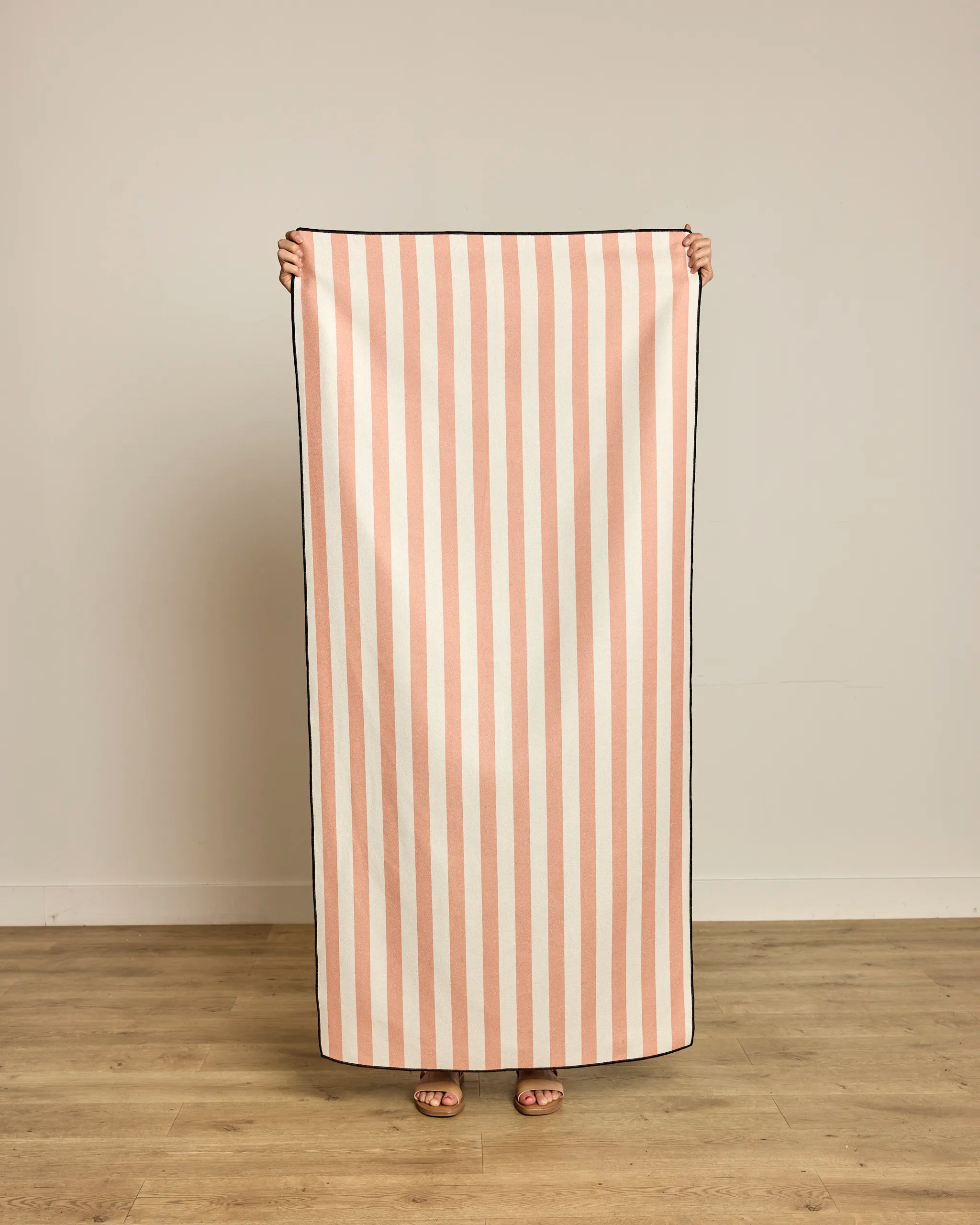 Indoors on a wooden floor, someone holds up Geometry’s Cabana Micro-loop Beach Towel, featuring vertical pink and white stripes. Made from recycled materials, this quick-drying towel covers their body except for their feet and hands.